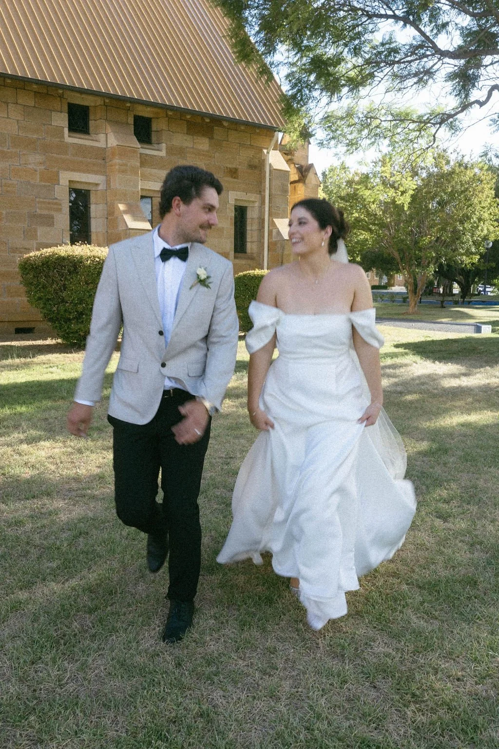 A bride and groom walking outdoors on wedding day, with the bride in a white off-shoulder wedding gown and the groom in a light gray suit with black bow tie, smiling at each other.