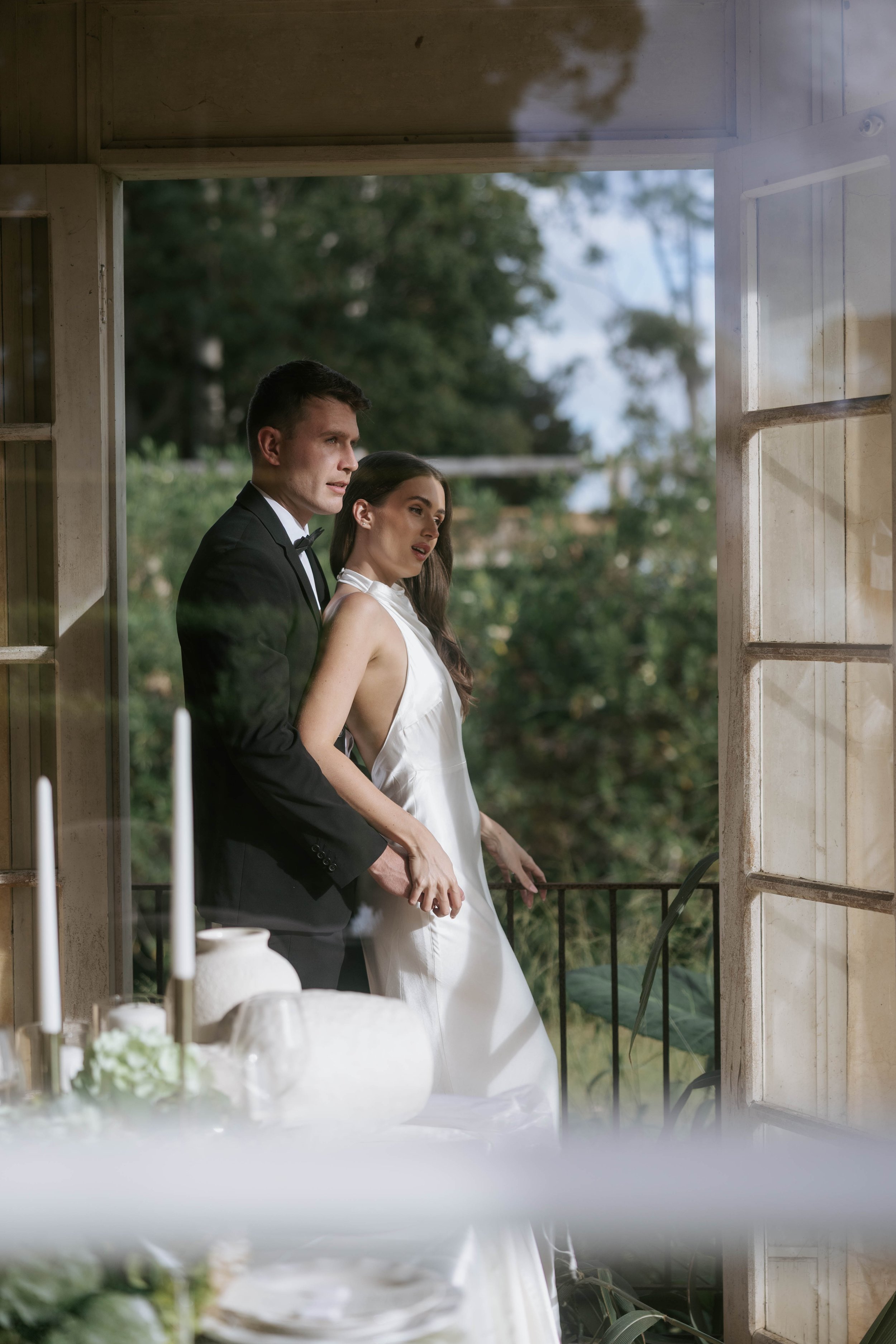A man in a tuxedo and a woman in a white dress are standing close together on a balcony, looking at something outside. The scene is outdoors with trees and sky in the background.