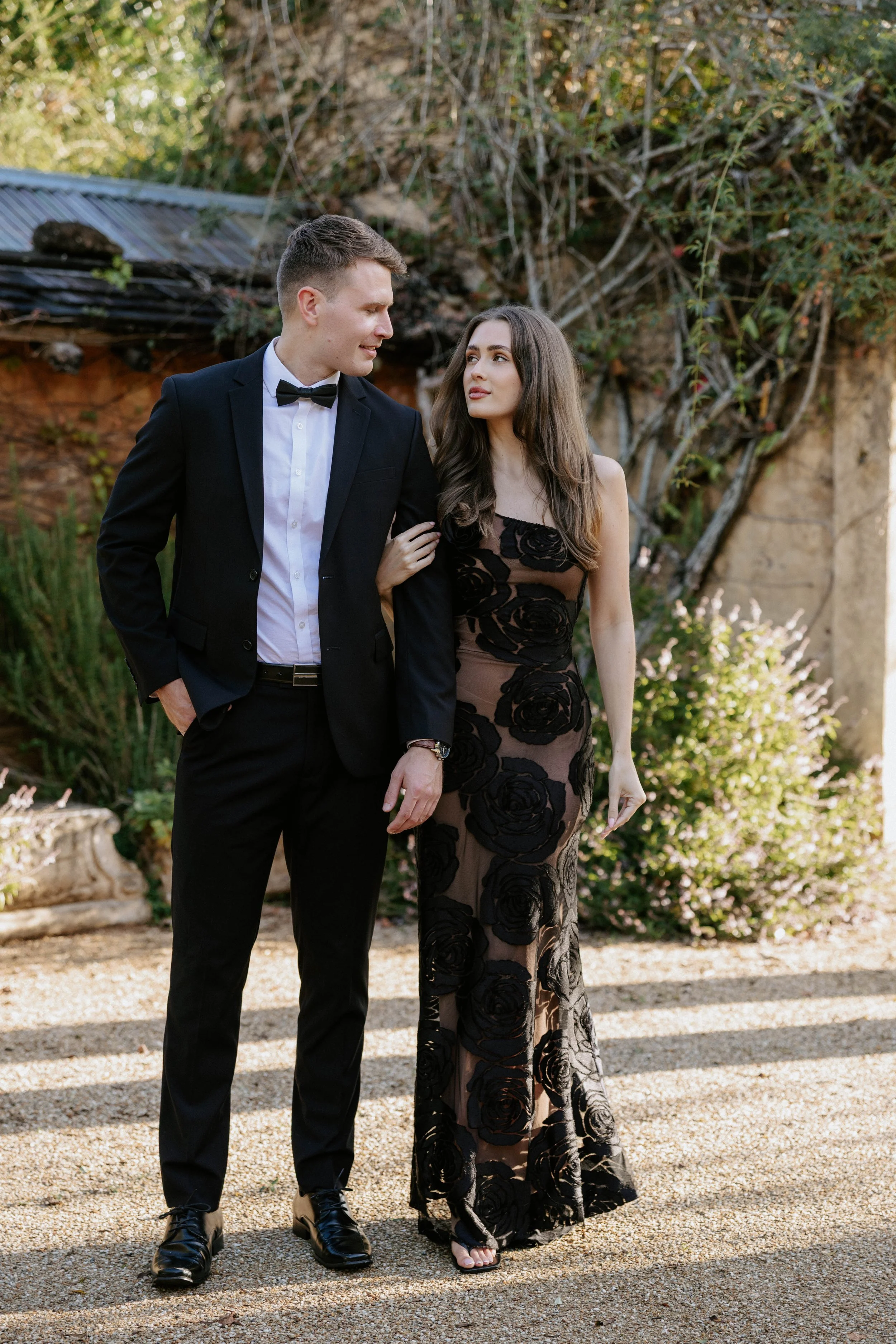 A young man and woman dressed elegantly walking outdoors in a garden setting. The man is in a black tuxedo with a bow tie, and the woman is in a black and nude floral lace gown. They are looking at each other with the man's arm linked with the woman's.