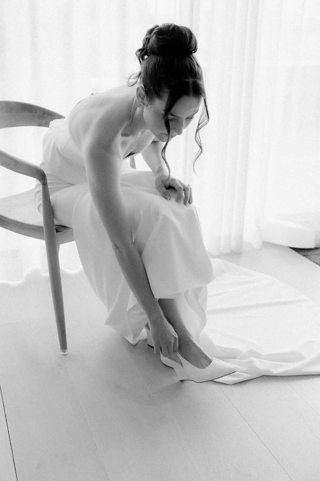 A woman in a strapless dress is sitting on a chair, putting on a white high-heeled shoe, with her hair styled in an elegant updo, in a bright, minimalistic room.