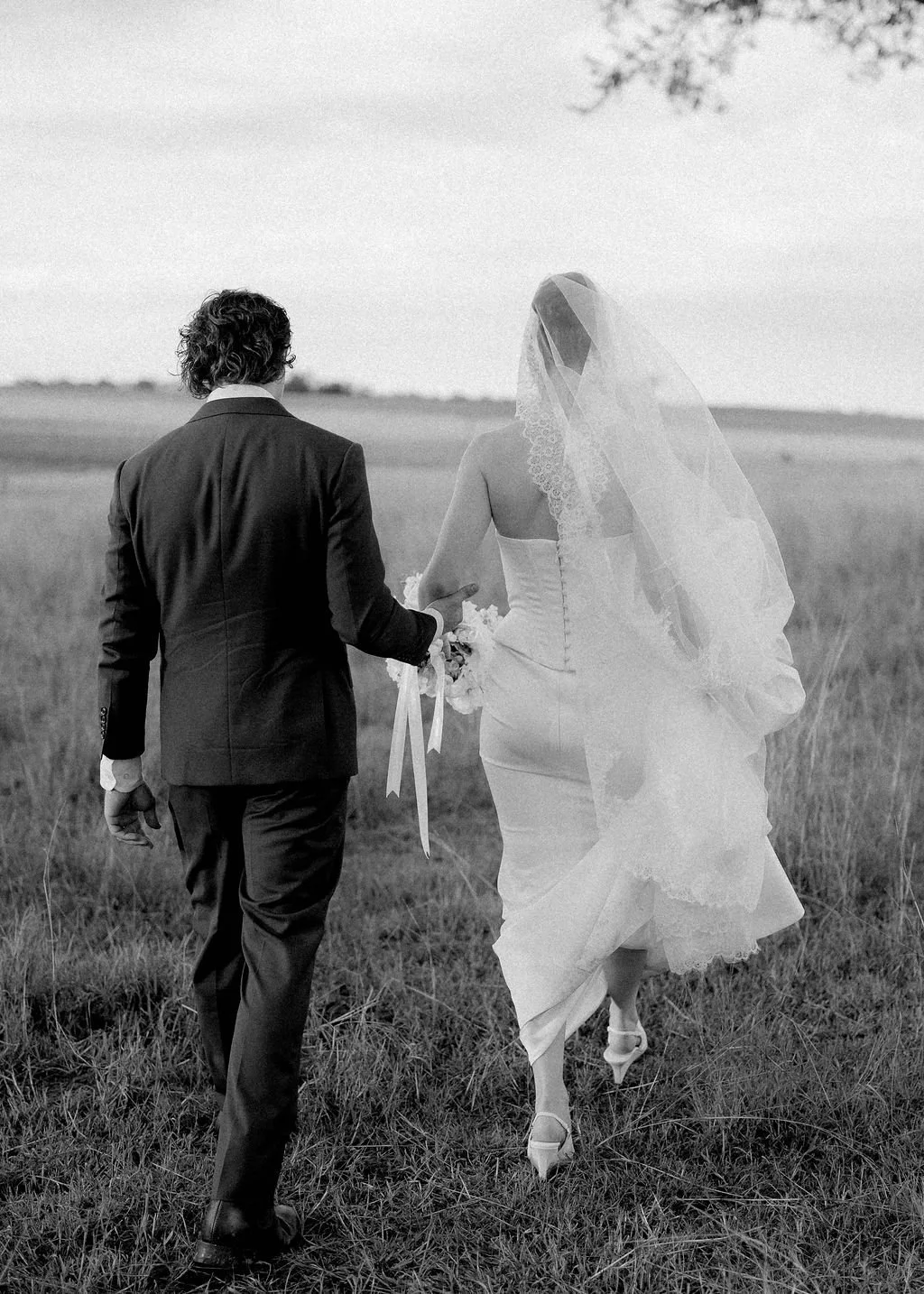 A black and white photo of a bride and groom walking away in an open field, with the bride wearing a wedding dress and veil and the groom in a suit.