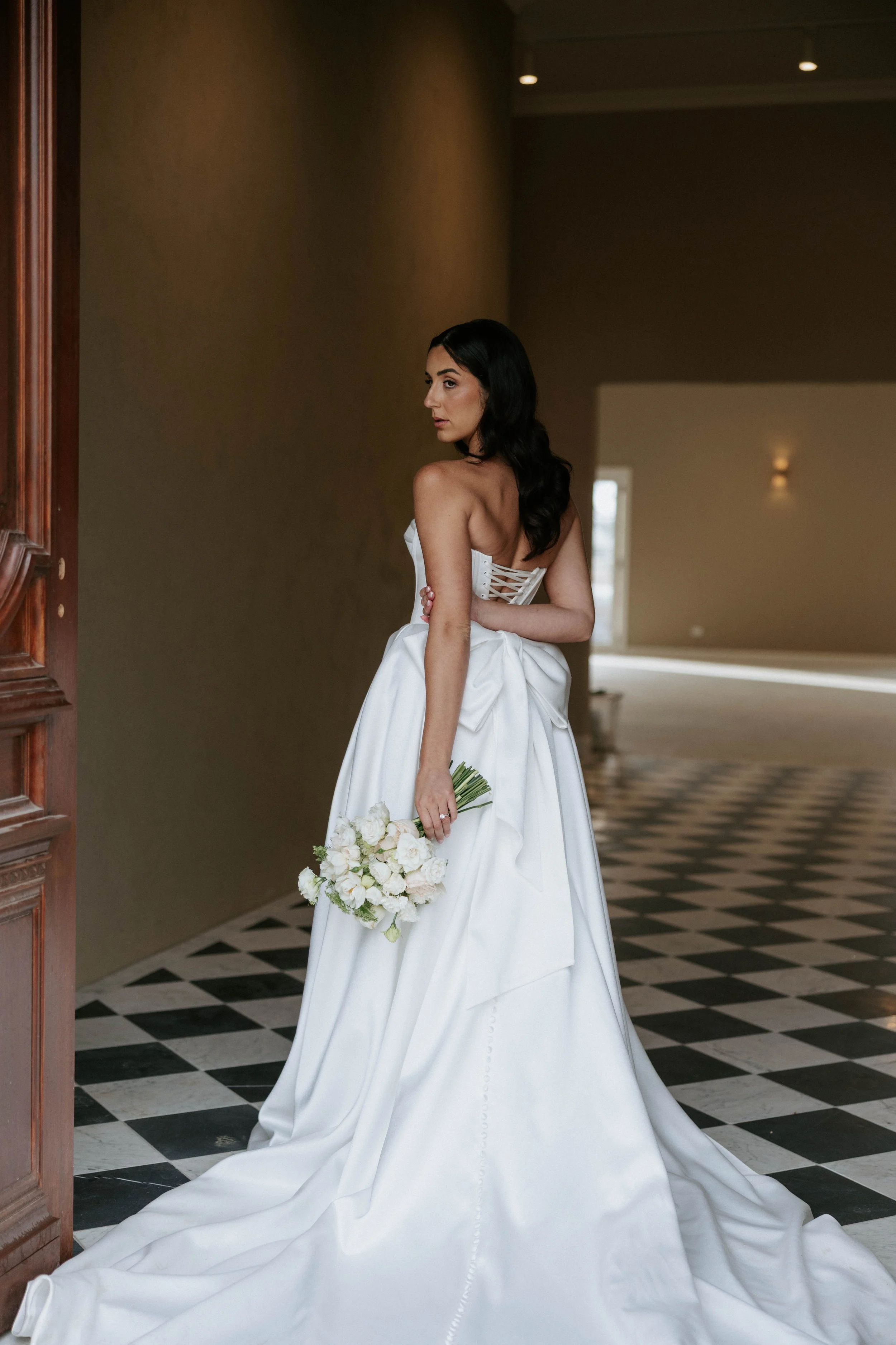 A woman in a white wedding dress holding a bouquet of white flowers standing in a hallway with checkered black-and-white tile flooring and dimly lit walls.
