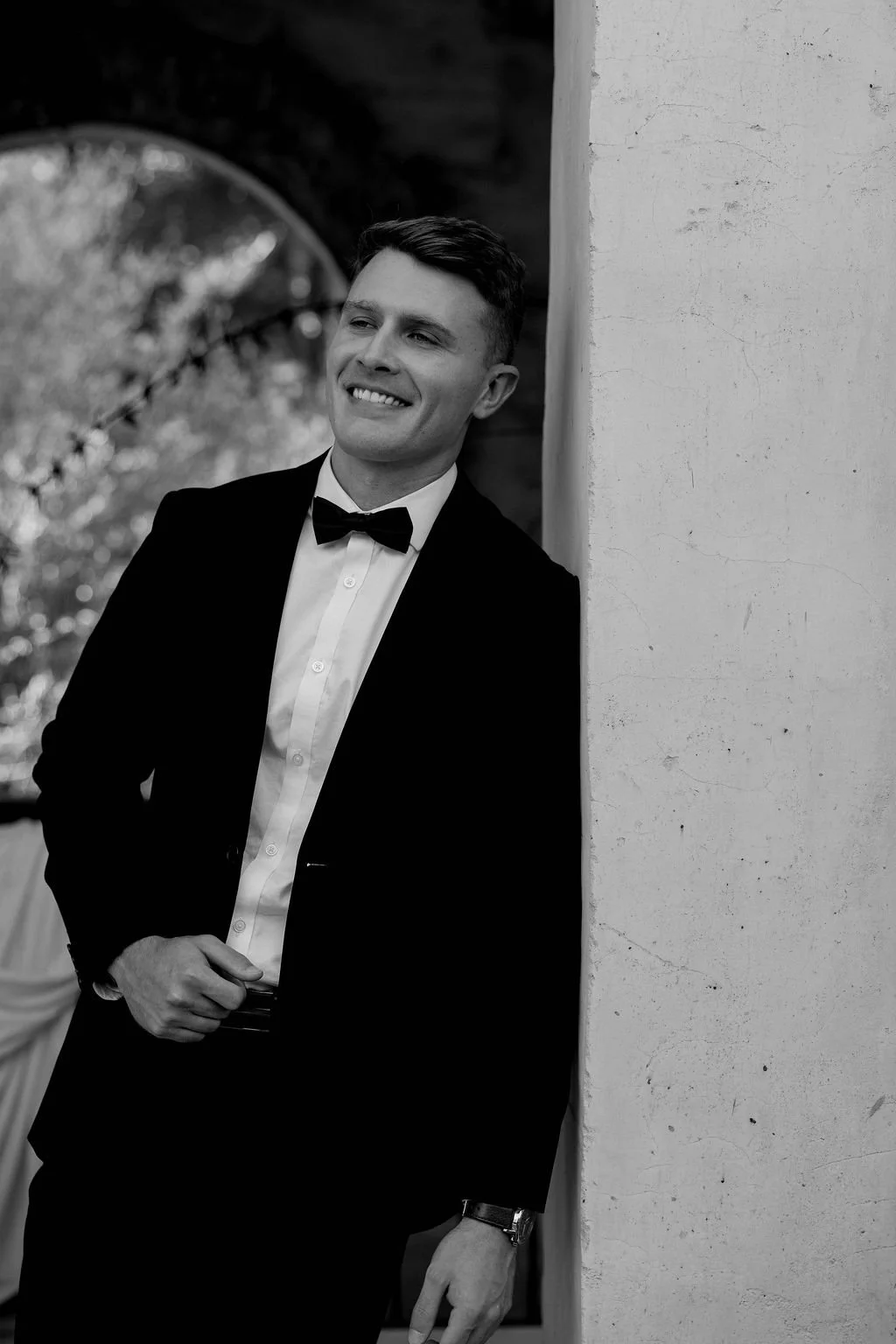A young man in a tuxedo with a bow tie, smiling, leaning against a white wall outdoors.