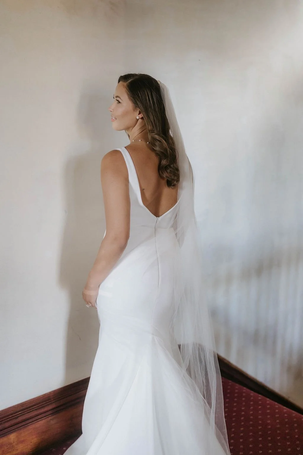 A bride in a white wedding gown with a deep V back and a long veil, smiling and looking to her left, standing indoors against a plain light-colored wall and dark wood floor.