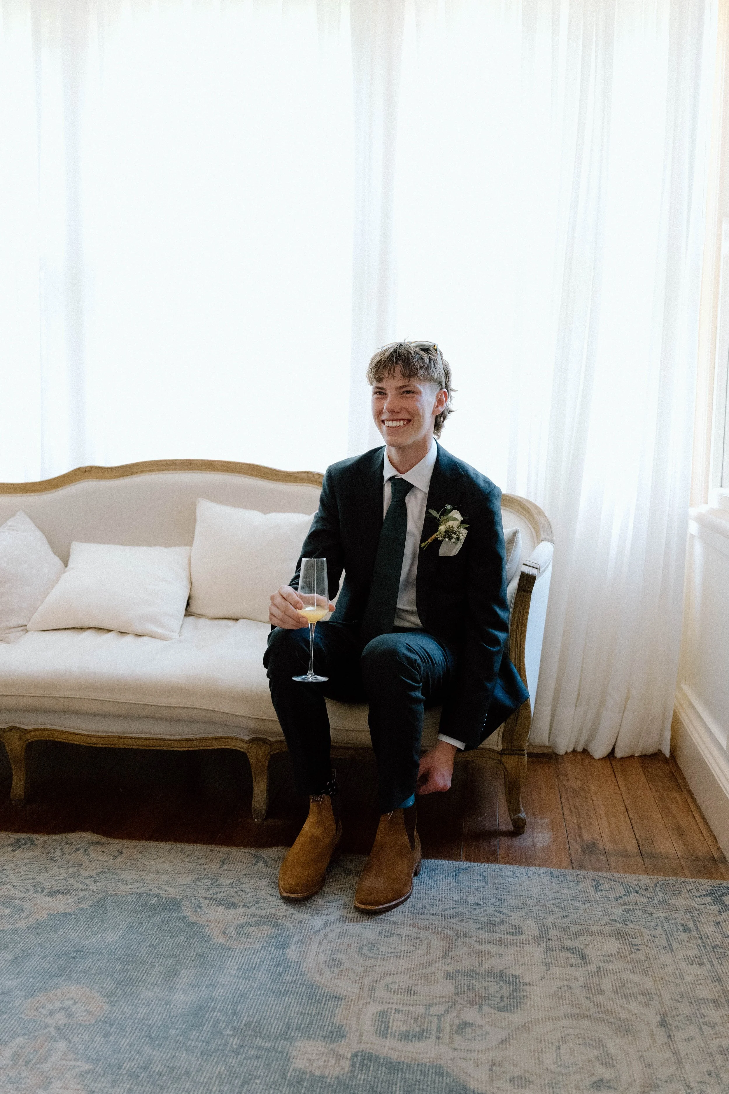 A young man in a tuxedo with a boutonniere is sitting on a vintage sofa, holding a champagne flute, smiling, wearing brown boots, in a bright room with white curtains and wooden floors.