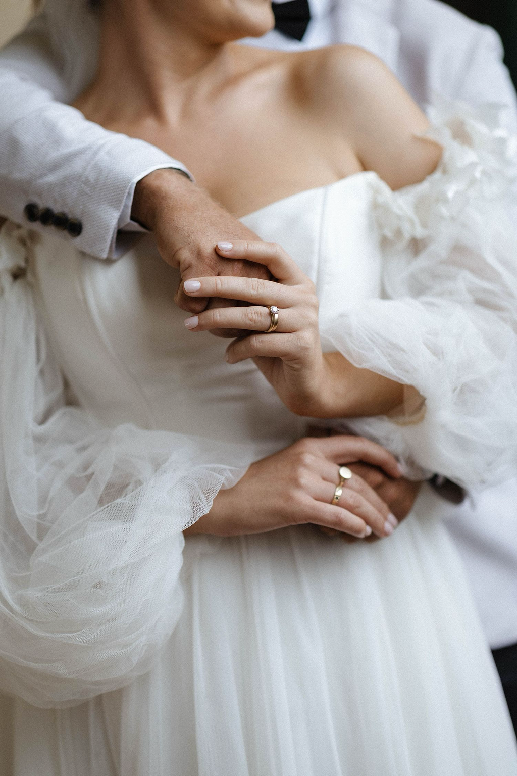 A bride and groom holding hands during their wedding, with wedding rings visible on their fingers, and the bride wearing an off-shoulder wedding dress and a veil.