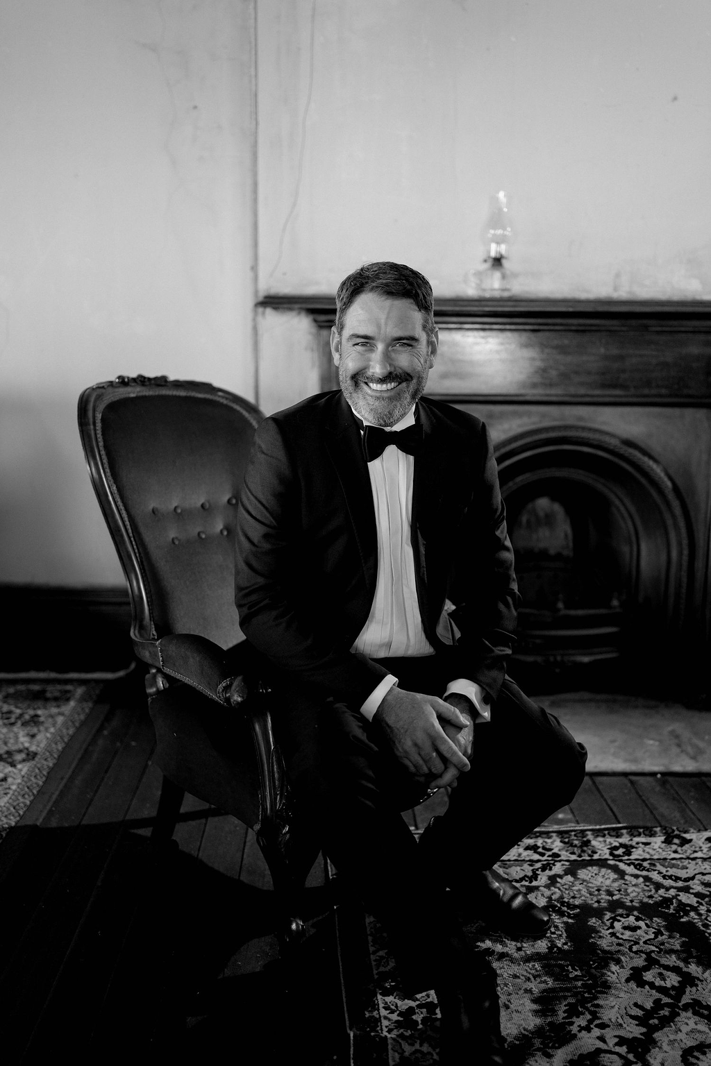 A man in a tuxedo sitting on a vintage armchair in a room with a fireplace, smiling.