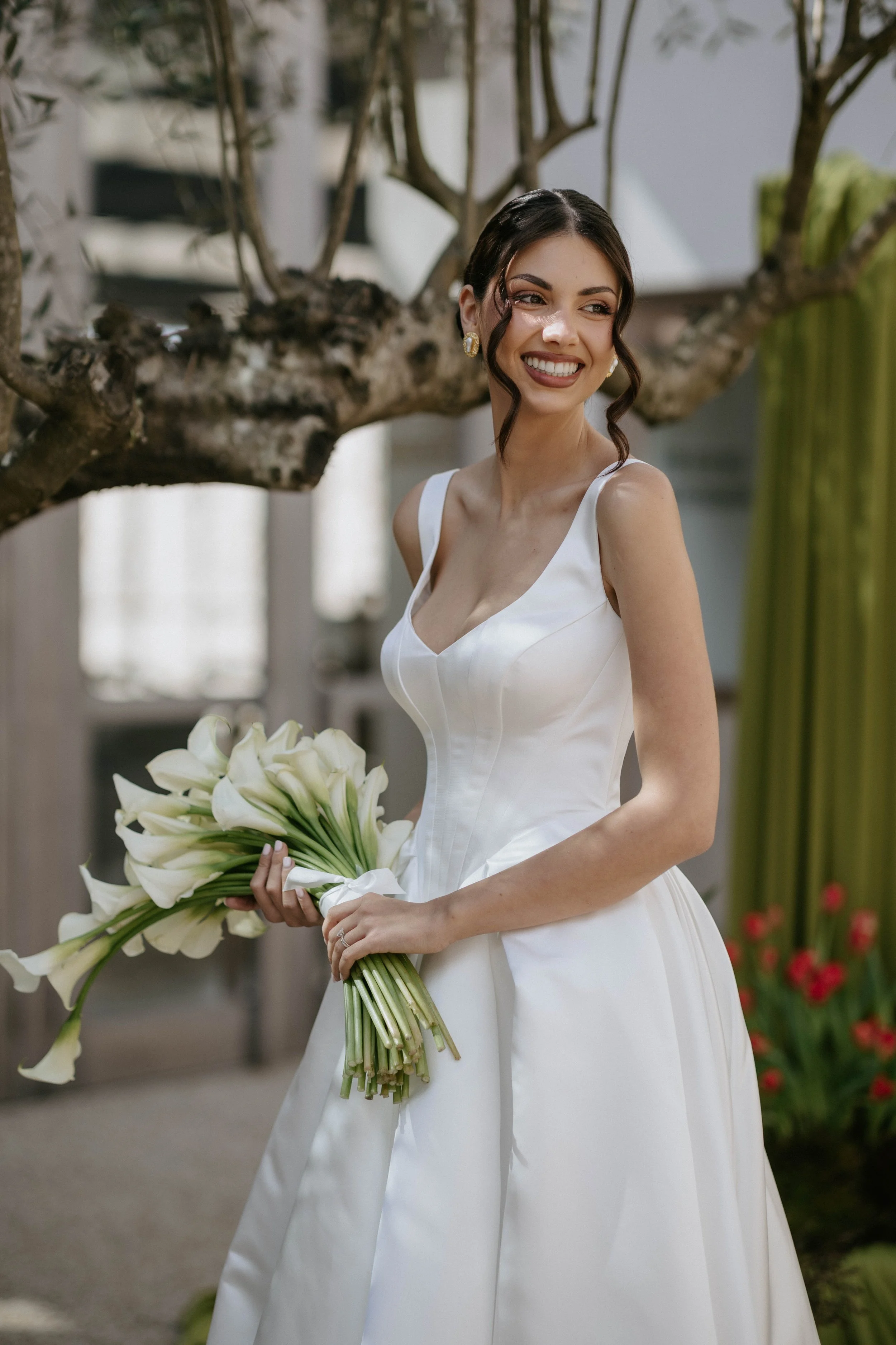 A woman in a white dress holding a bouquet of white calla lilies, smiling and standing outdoors near a tree and a building.