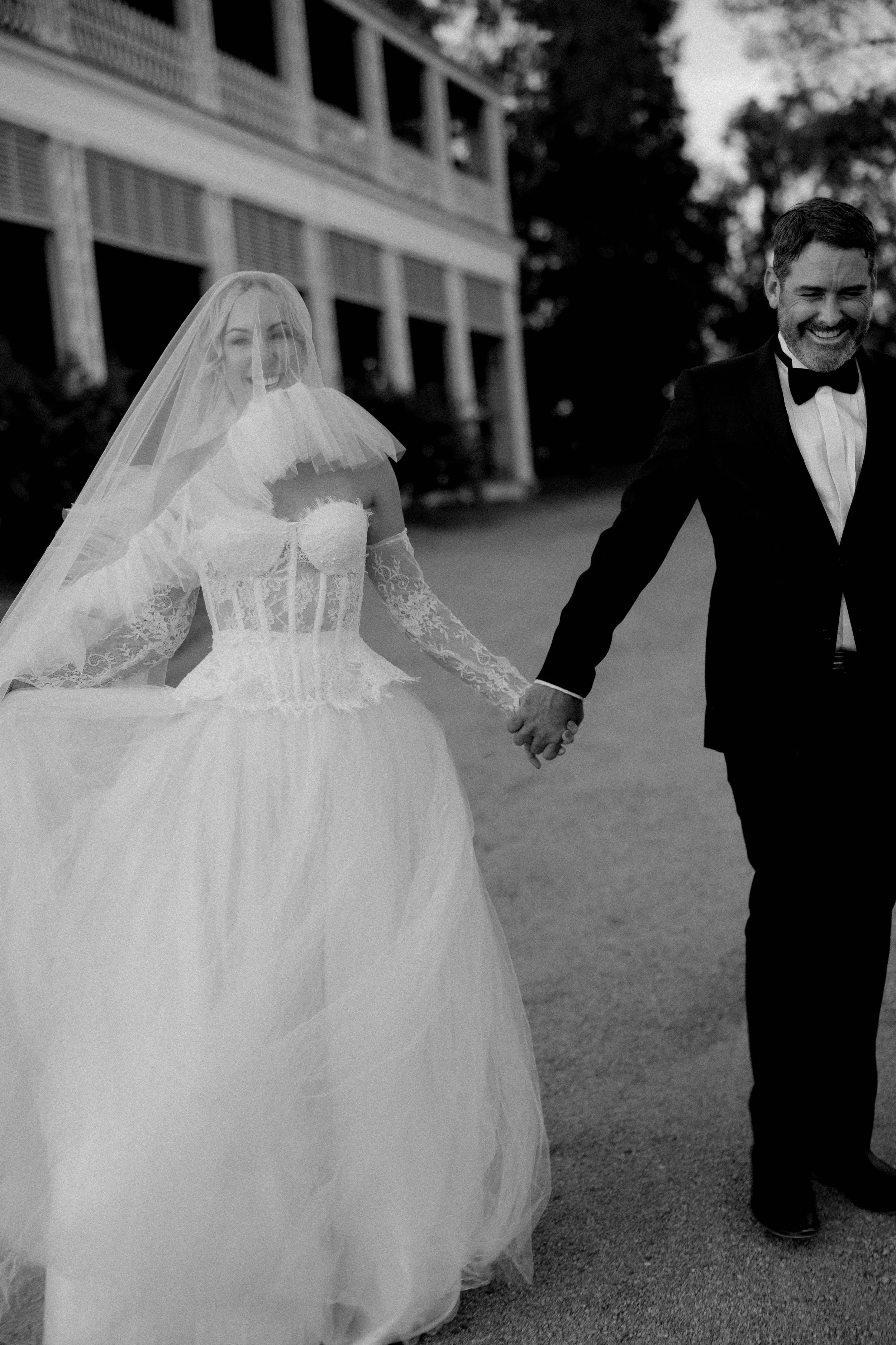 A bride in a wedding dress and veil holding hands with a groom in a tuxedo, smiling outdoors.