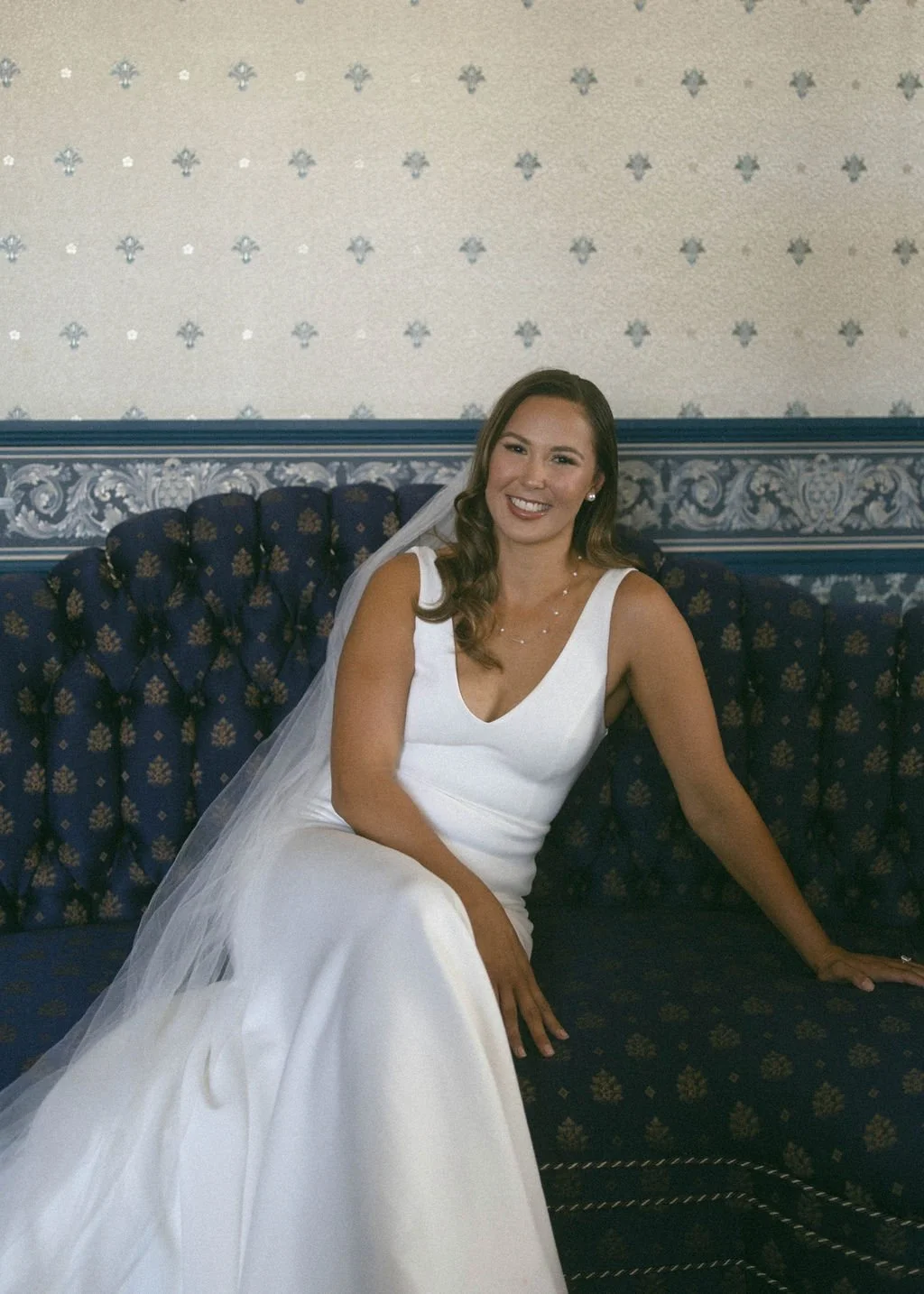 A woman in a white wedding dress sitting on a dark blue patterned sofa, smiling, with a veil and jewelry, in an ornately decorated room.