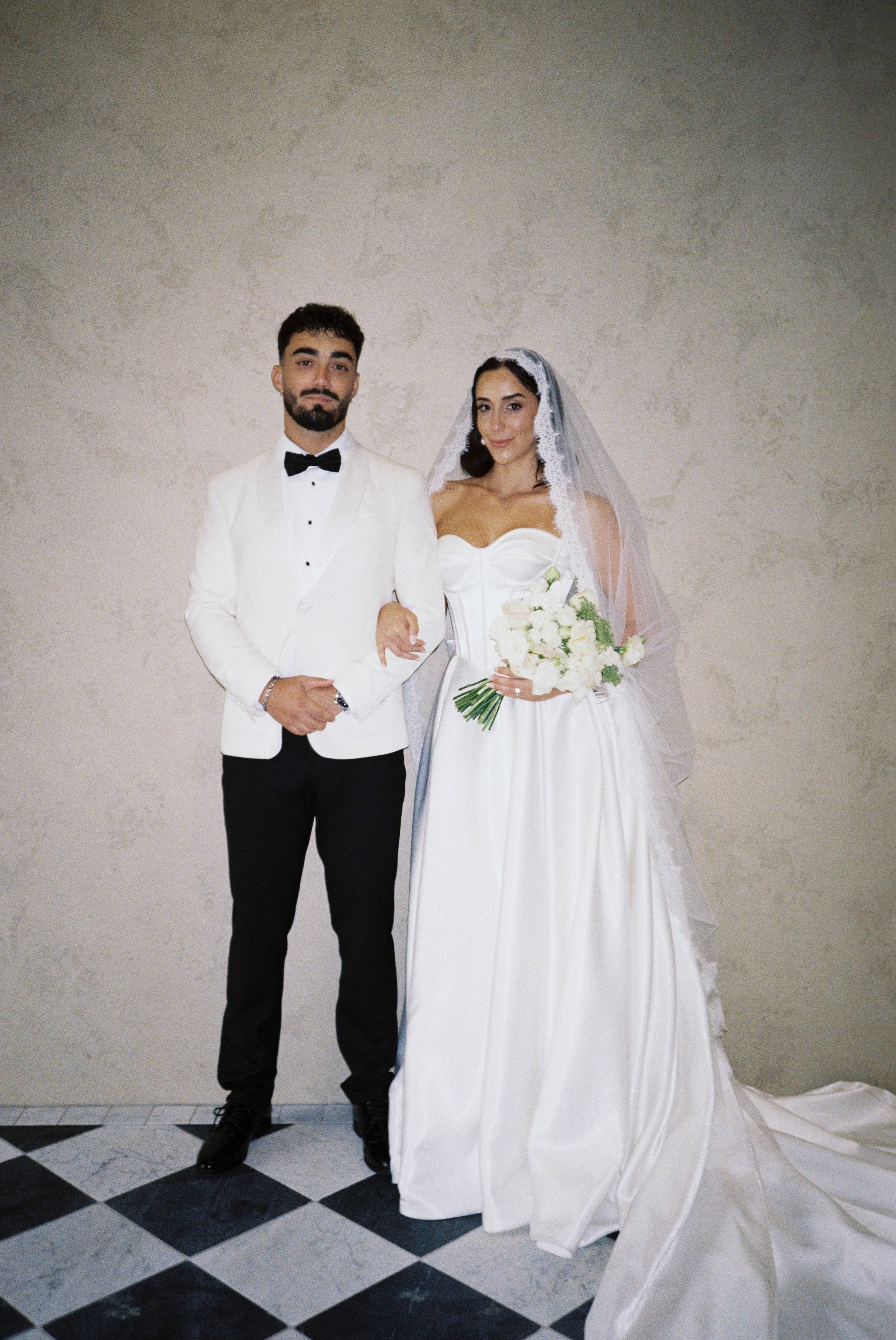 A bride and groom in wedding attire standing indoors against a plain beige wall. The groom is wearing a white tuxedo jacket, black pants, black shoes, and a black bow tie. The bride is wearing a strapless white wedding gown, holding a bouquet of white flowers, and has a lace veil covering her dark hair.