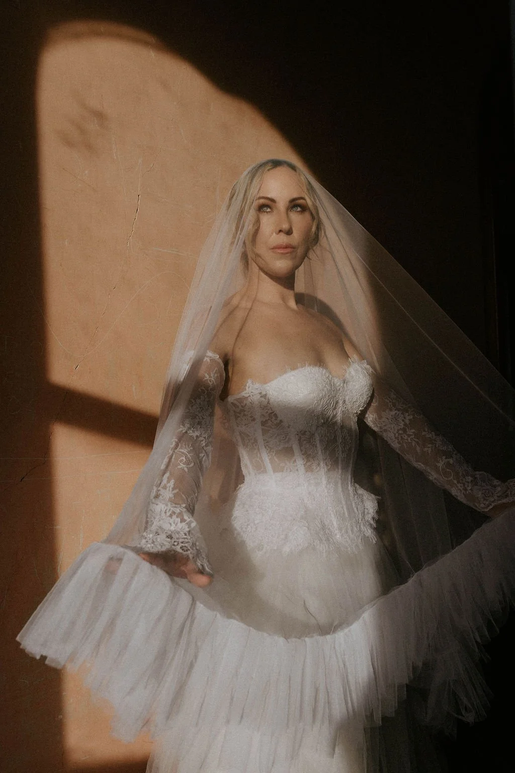 A woman in a wedding dress with lace details, standing in front of a warm-colored wall during a photoshoot.