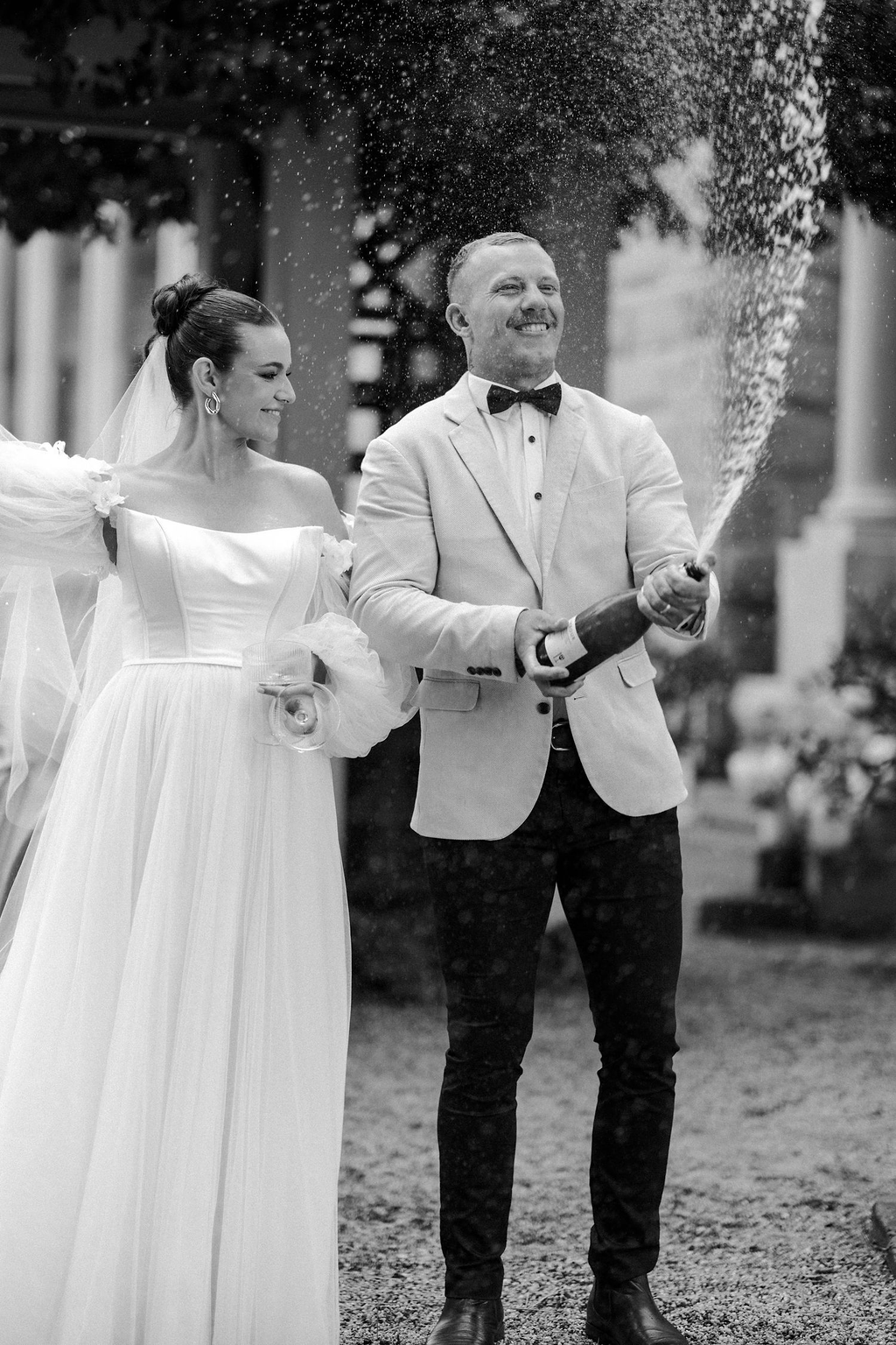 A newlywed couple celebrating their wedding outdoors with champagne, the groom is spraying champagne while the bride holds a glass