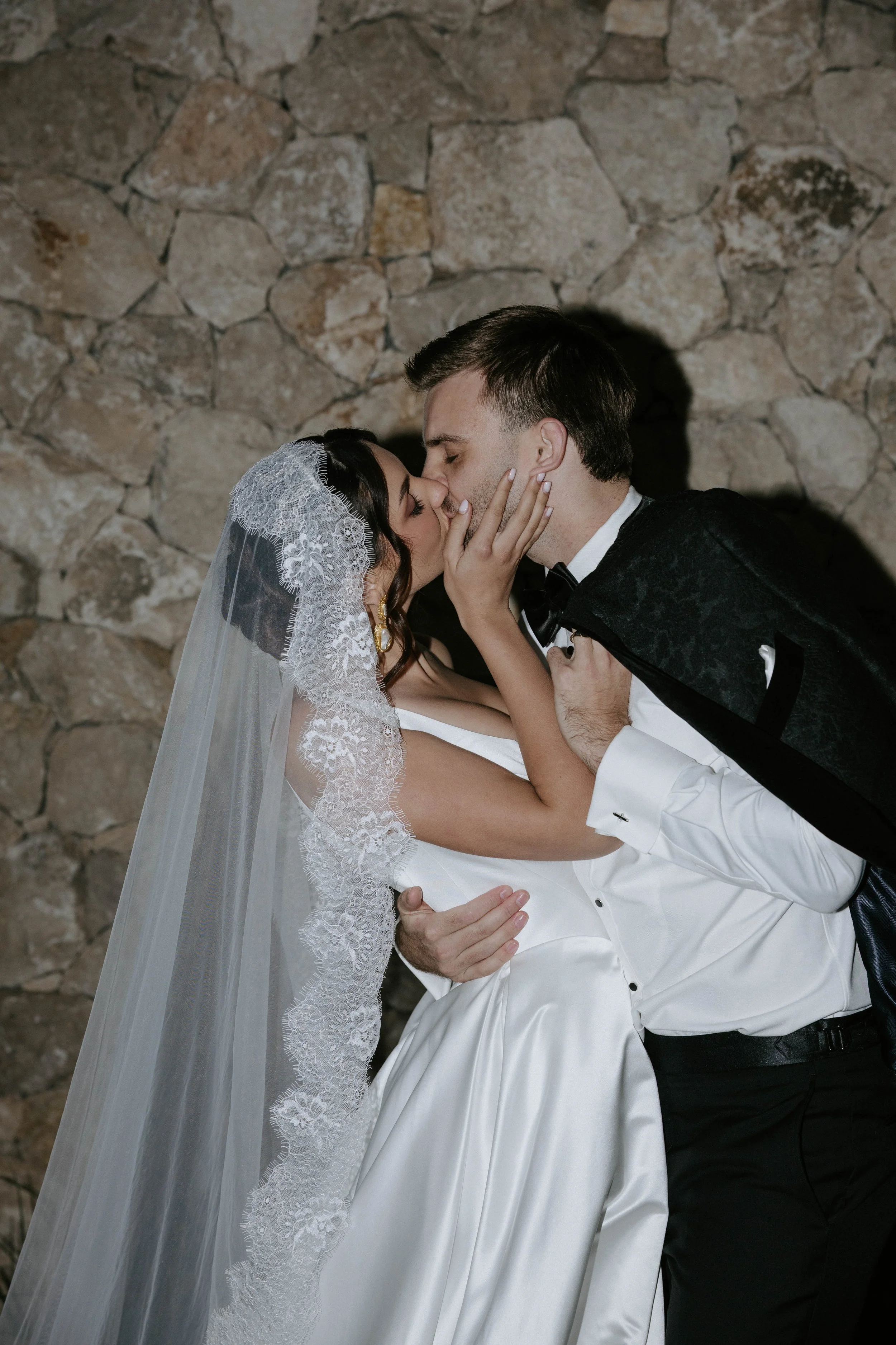 A bride and groom kissing, with the bride wearing a lace wedding veil and dress, and the groom in a tuxedo, in front of a stone wall.