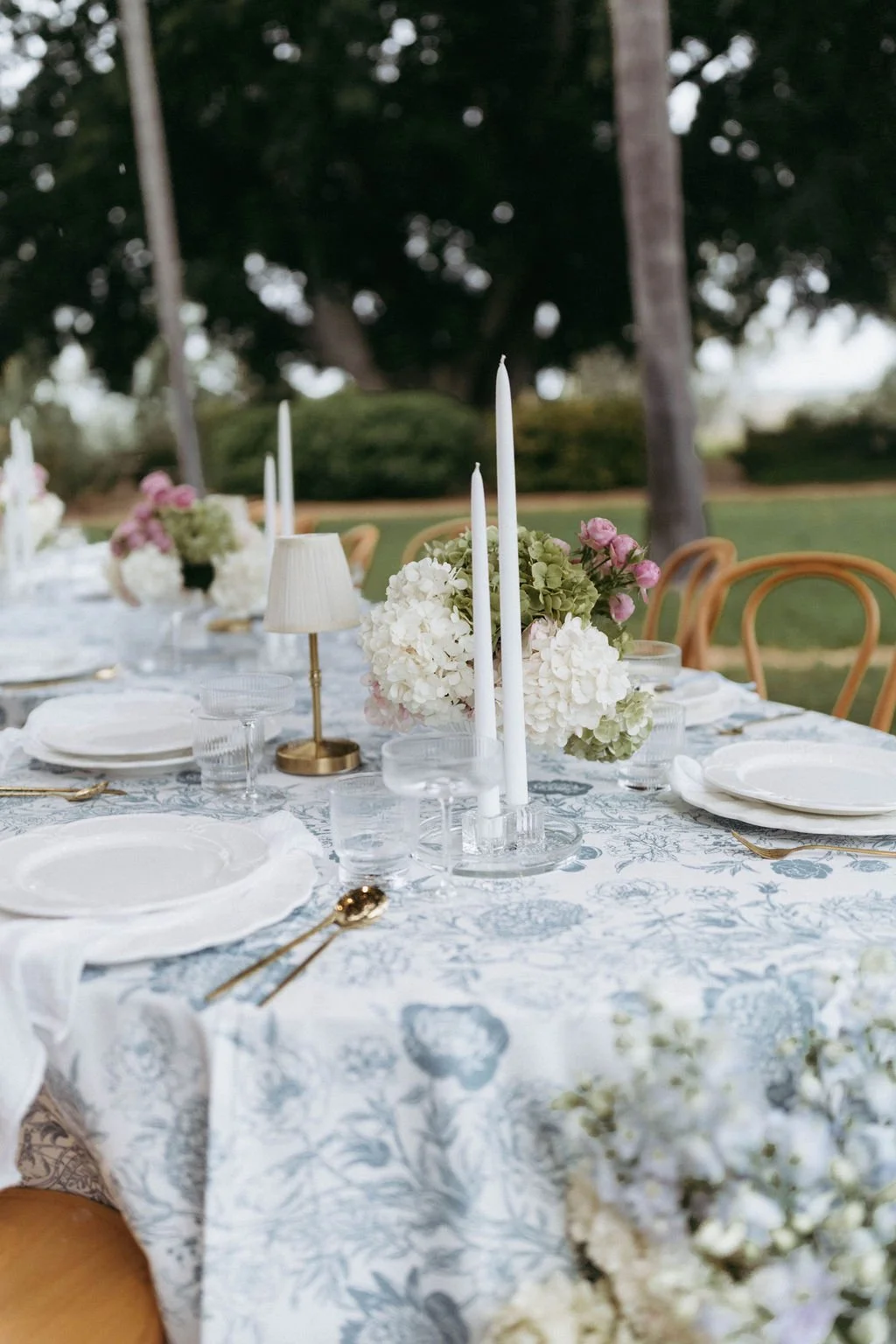 A beautifully set outdoor dining table with white plates, gold flatware, glassware, and a floral centerpiece with white, pink, and green flowers. Candles and small lamps decorate the table, with trees and a grassy area in the background.