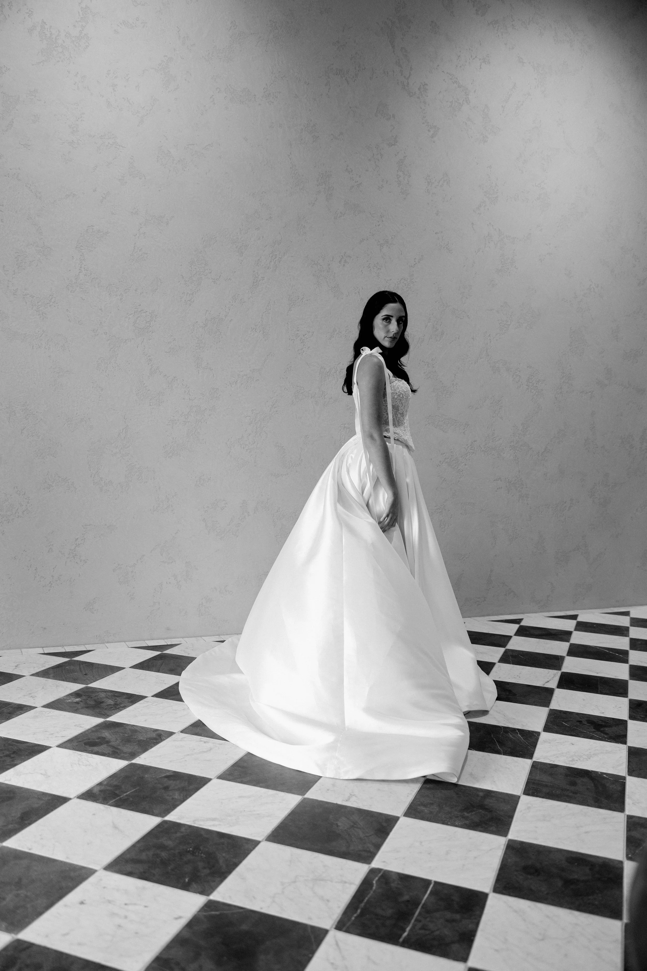A woman in a long, flowing white gown standing in a room with a checkered black-and-white floor and a plain wall background.