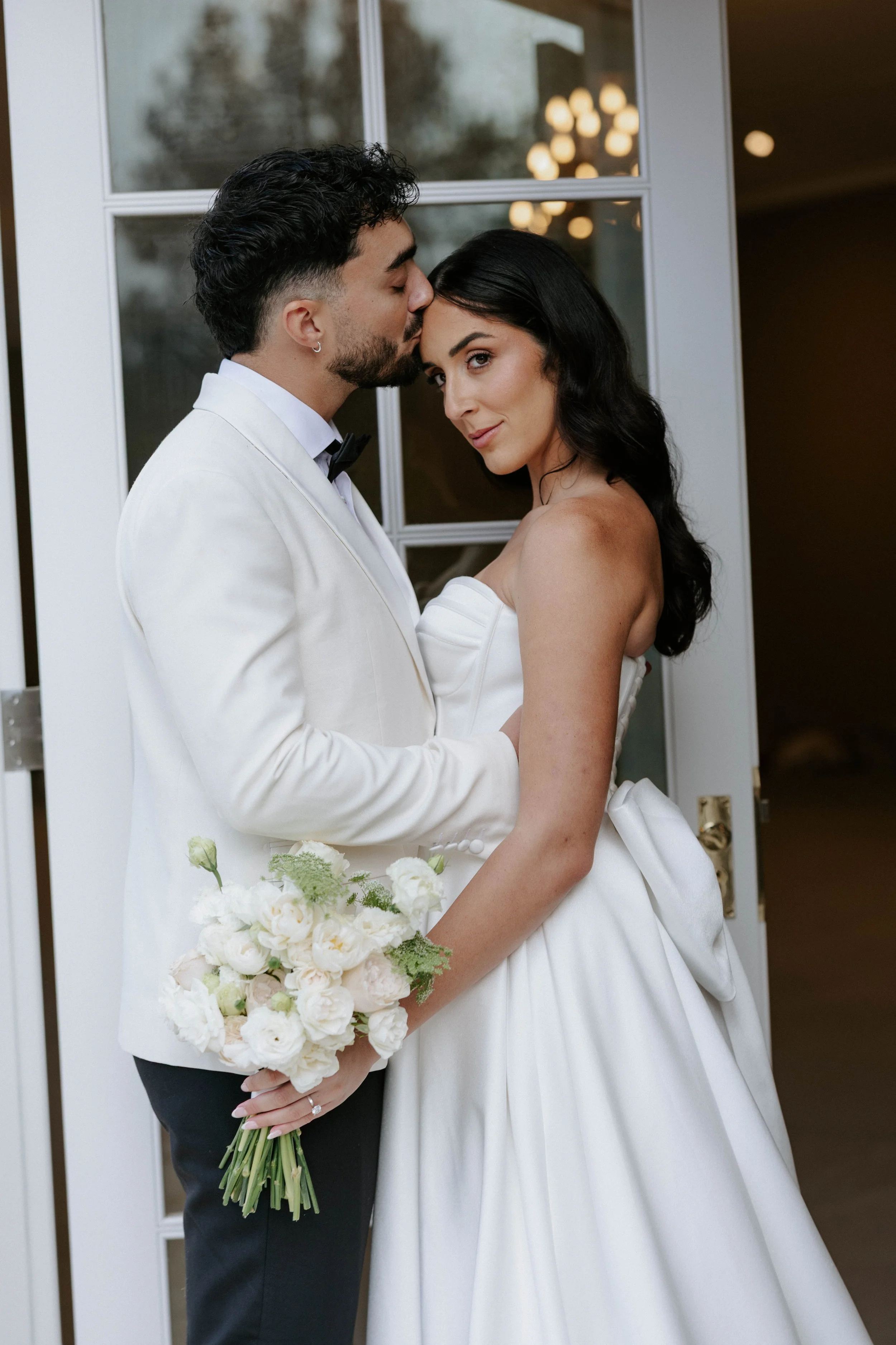 A newlywed couple standing close together, with the groom kissing the bride's forehead. The bride is holding a bouquet of white flowers and wearing an off-shoulder wedding dress. They are standing indoors near a window, with warm lighting in the back