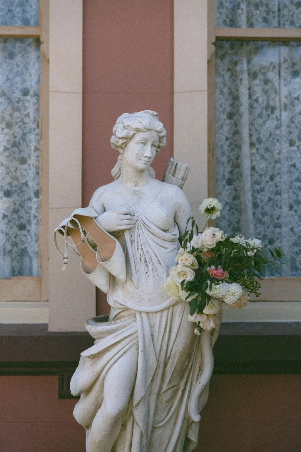 A statue of a woman holding a bouquet of flowers, with a gray high-heeled shoe and other items hanging from her arm, set against a building with windows.