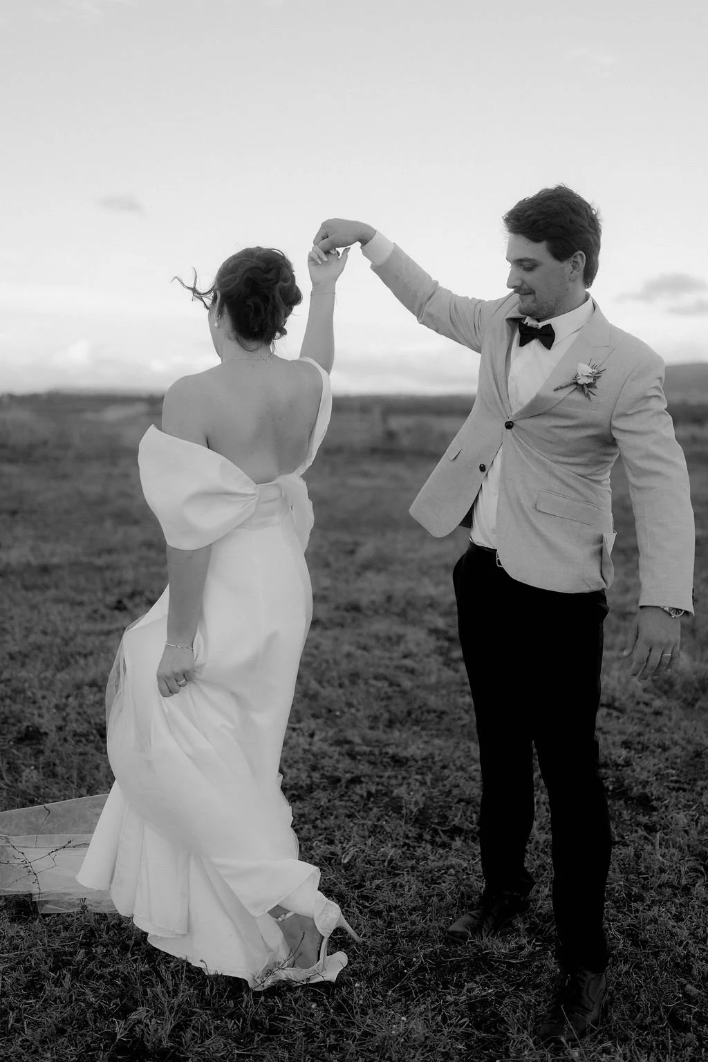 A black-and-white photo of a bride and groom dancing in a field outdoors during sunset or sunrise, with the groom holding the bride's hand.