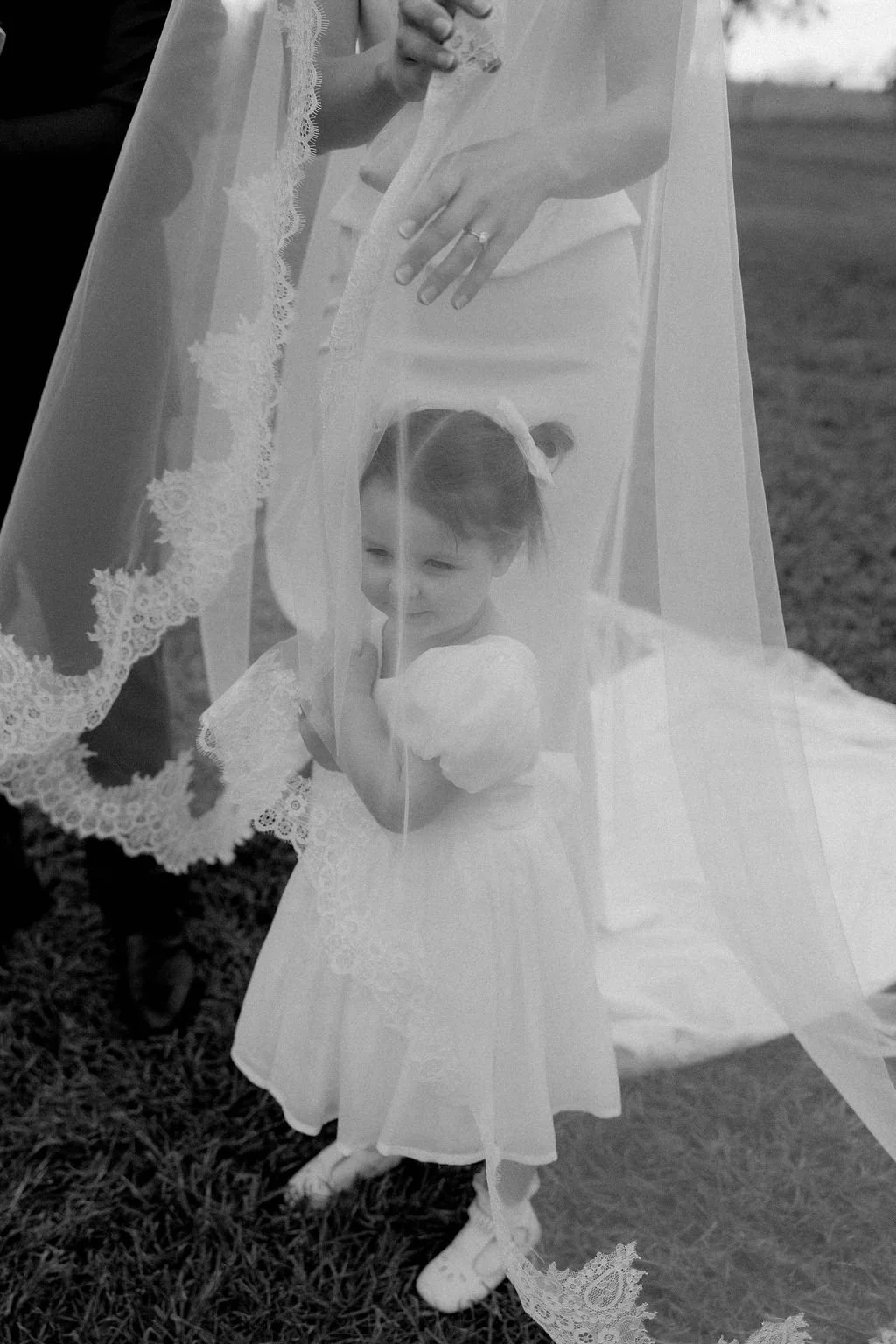 A young girl dressed in a white dress is standing outdoors under a lace veil, looking through it with a shy smile. An adult's hands are adjusting the veil, which has lace trim, and the scene appears to be a special occasion like a wedding or a formal