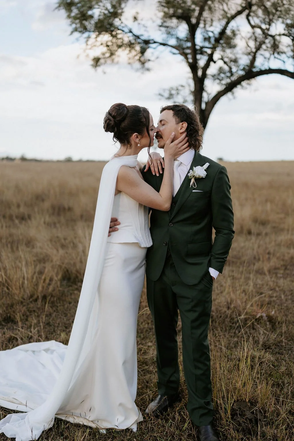 A bride and groom in a field, standing close with faces touching, embracing during their wedding photos. The bride has her hand on the groom's face and is wearing a white dress with a cape, while the groom is dressed in a dark green suit with a bouto