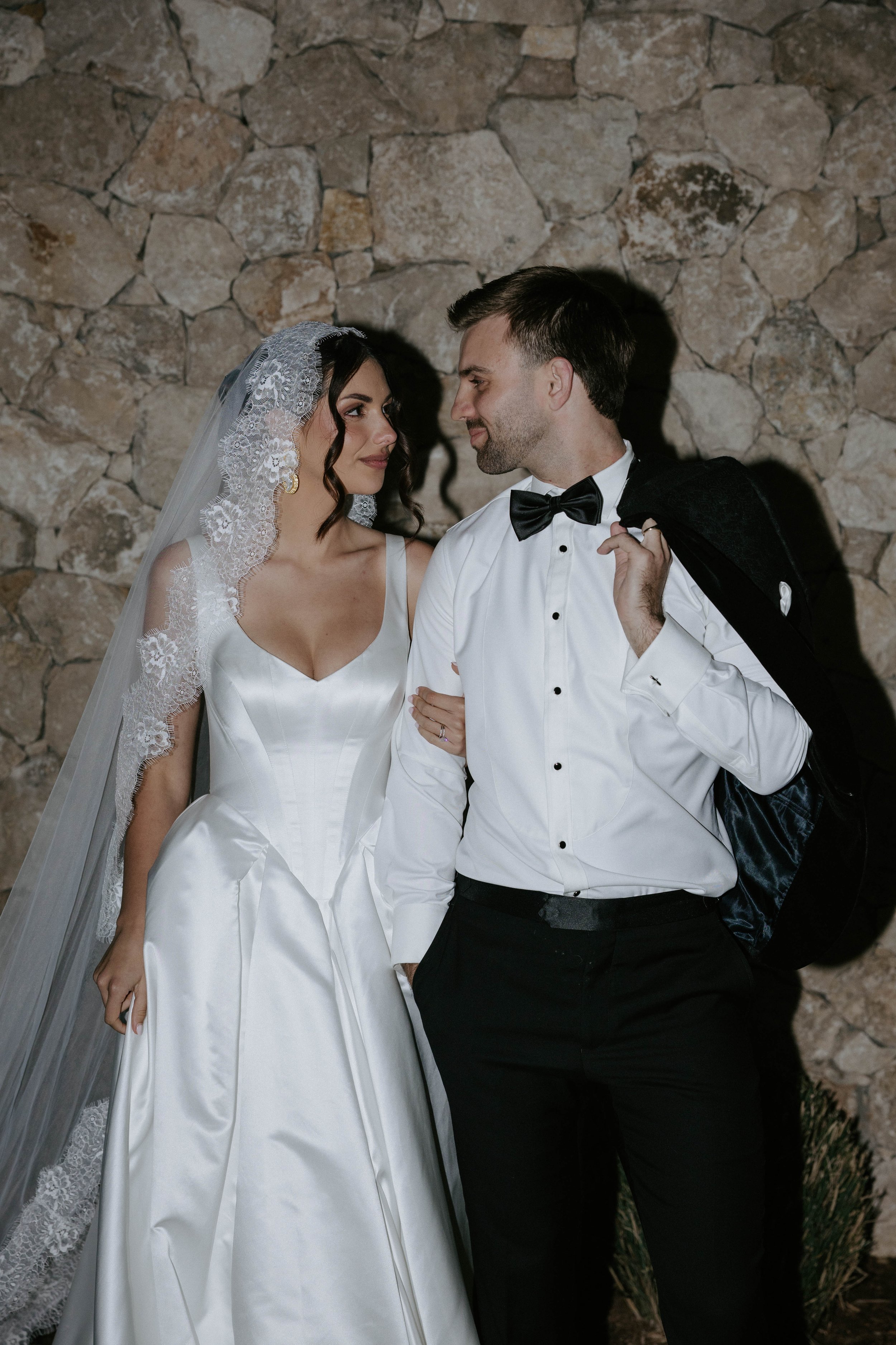 A bride and groom looking at each other in front of a stone wall. The bride wears a white satin wedding dress with lace detailing on her veil, and has dark, wavy hair. The groom wears a white tuxedo shirt with a black bow tie, and is holding a black 