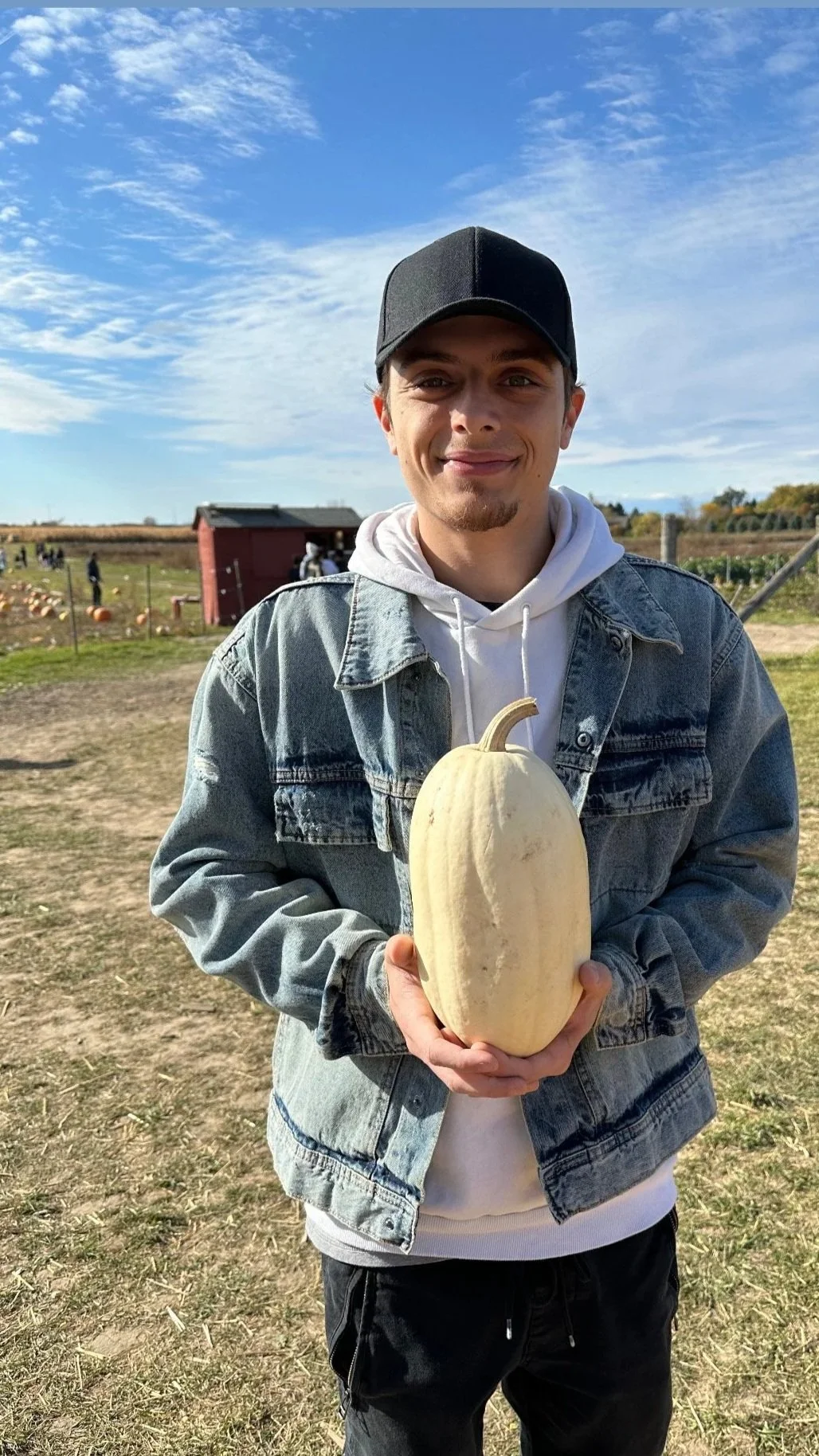 A young man holding a white pumpkin at a pumpkin patch on a sunny day, with blue sky and clouds in the background.