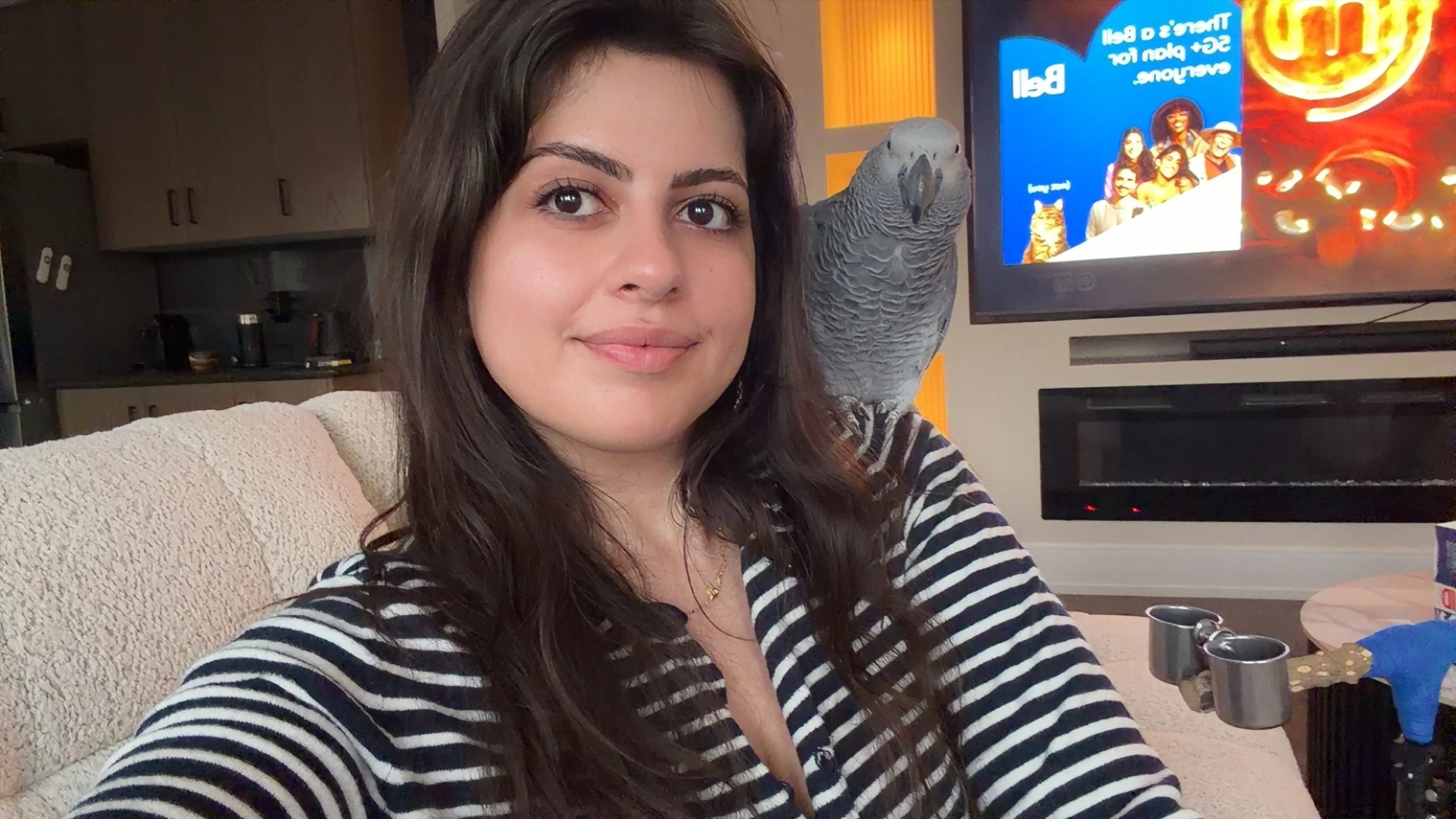 A woman with long dark hair and wearing a striped shirt takes a selfie indoors, with a television screen displaying colorful images and a parrot perched on her shoulder.