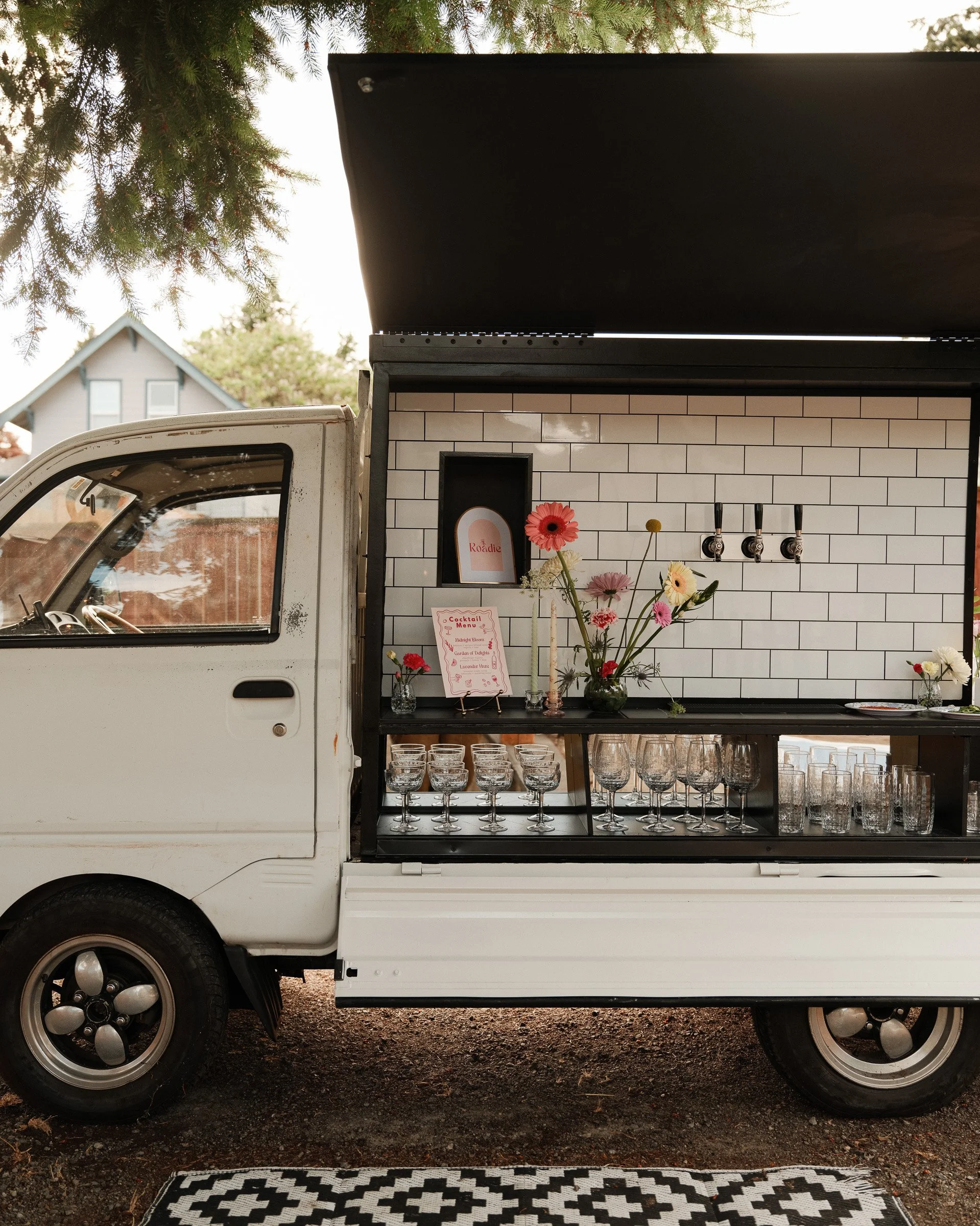 A food truck with a white brick wall design, displaying glasses and flowers inside, parked outside under a tree with a house in the background.