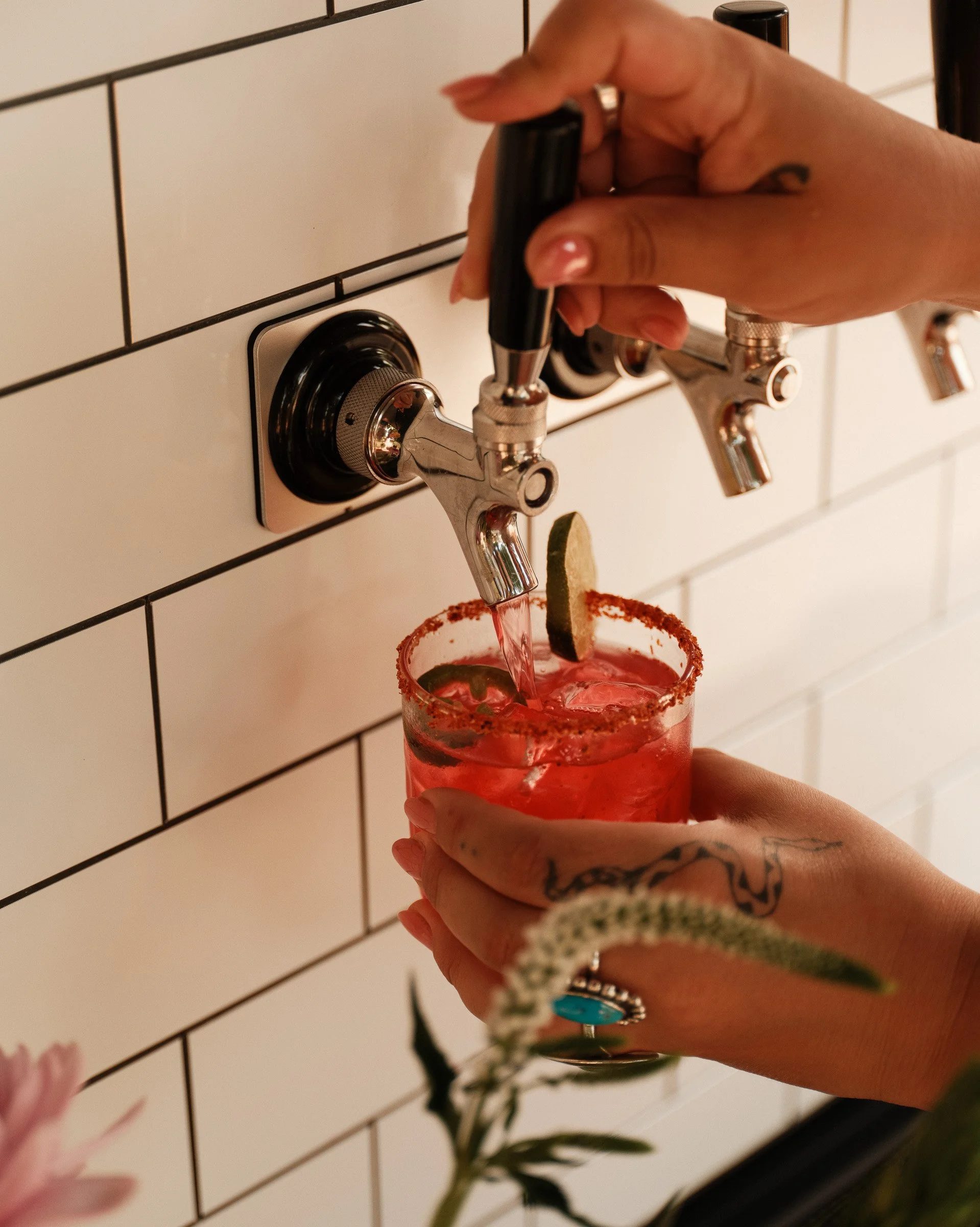 A hand filling a glass with pink cocktail from a faucet with a white tiled wall in the background.
