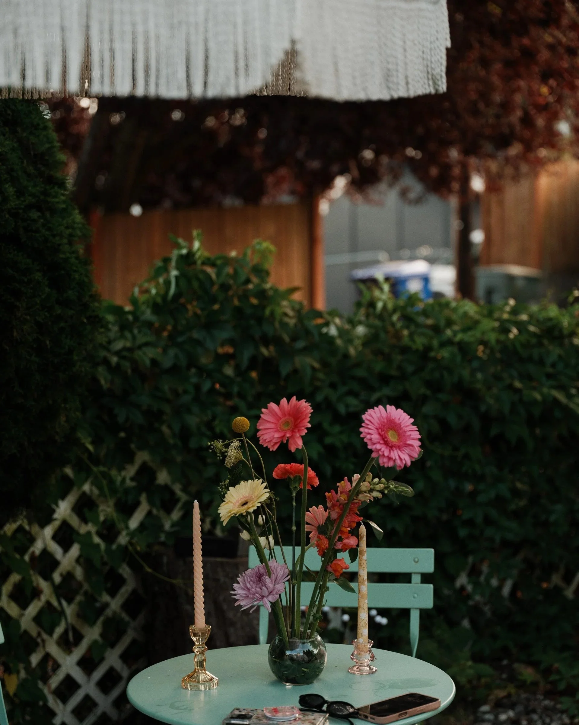 A round, mint green table with a small, clear glass vase holding pink, purple, and orange flowers, surrounded by three candles in decorative holders. There are various items including a pair of sunglasses, a smartphone, and small containers on the ta