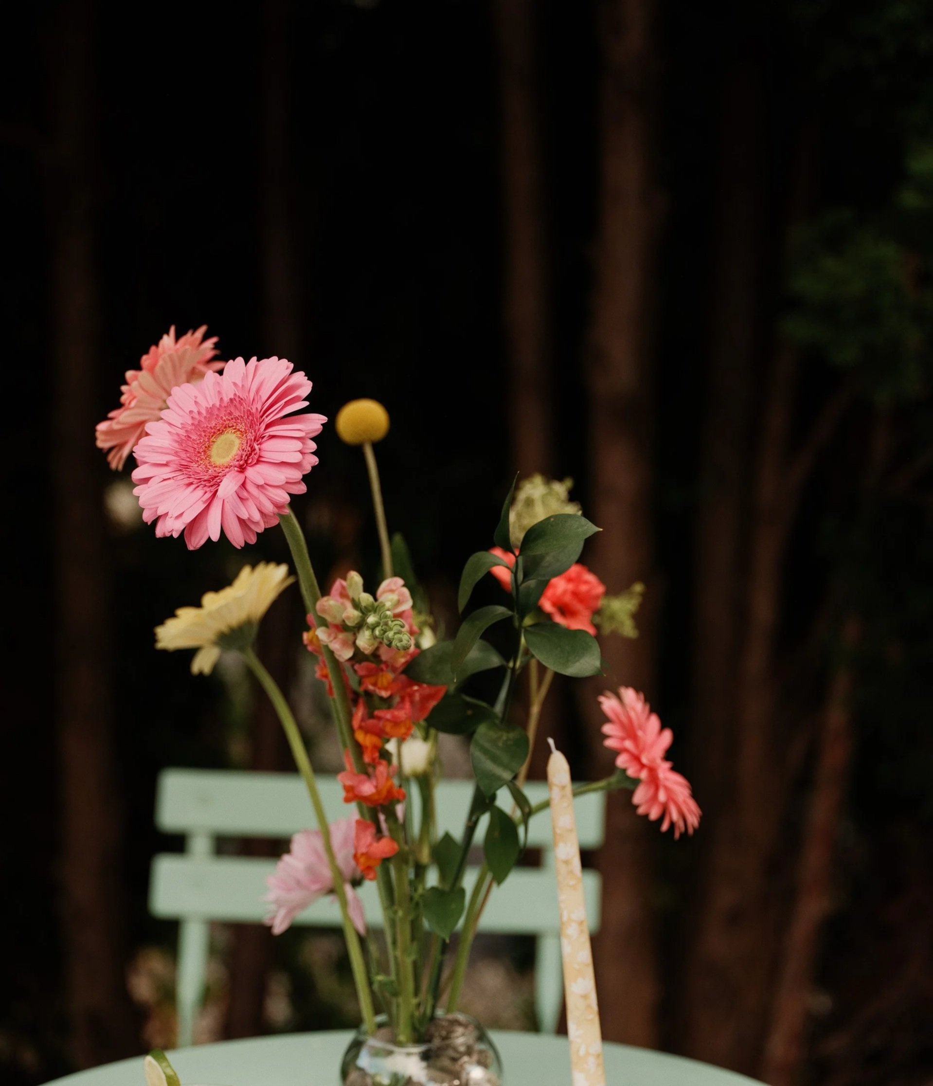 Arrangement of pink, yellow, and orange flowers in a glass vase on a green table with a black background.