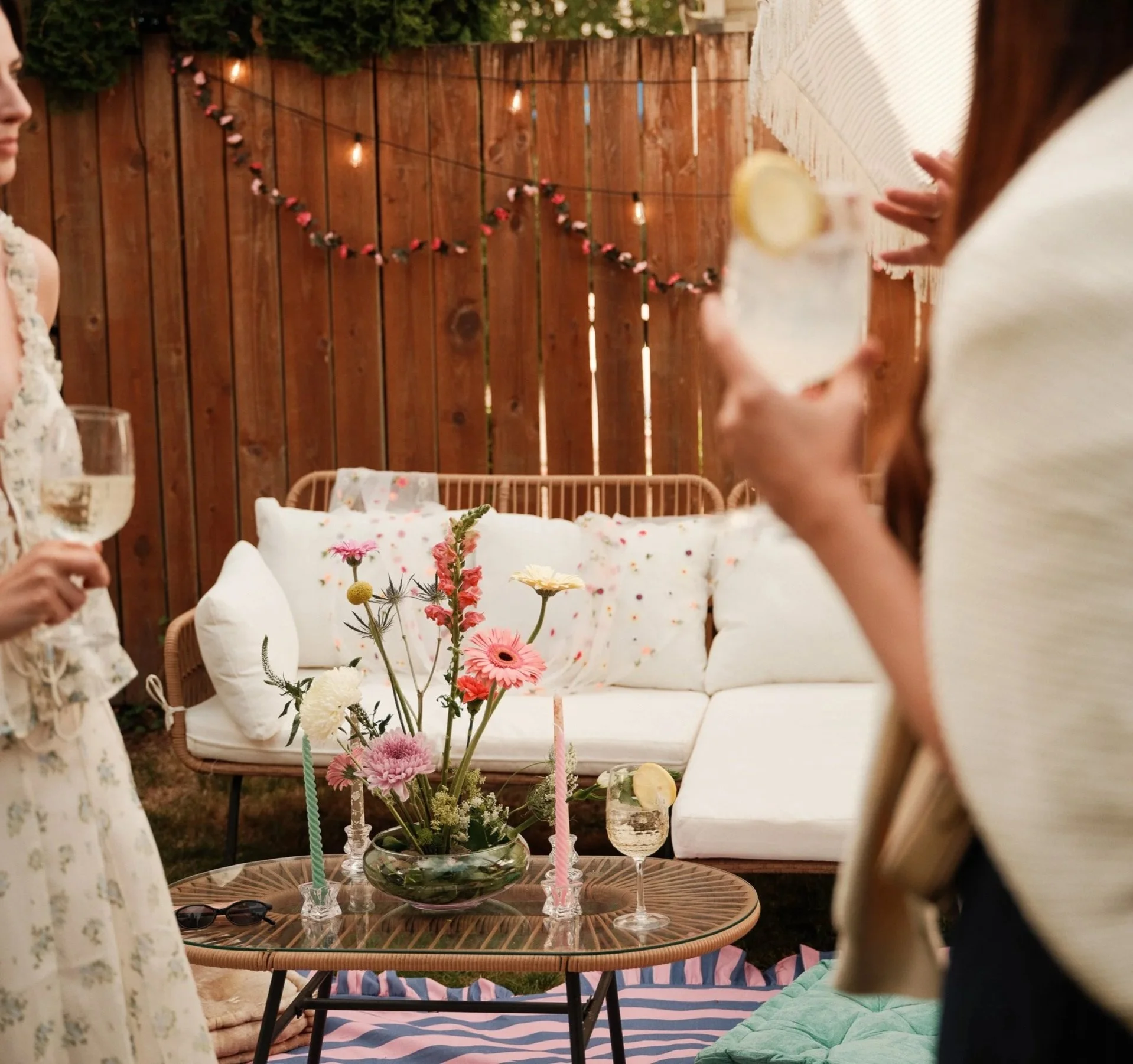 A backyard party scene with a table holding a floral centerpiece, glassware with lemon slices, and candles. There is a white outdoor sofa with cushions, a striped rug, and string lights on a wooden fence in the background. Two women are partially vis