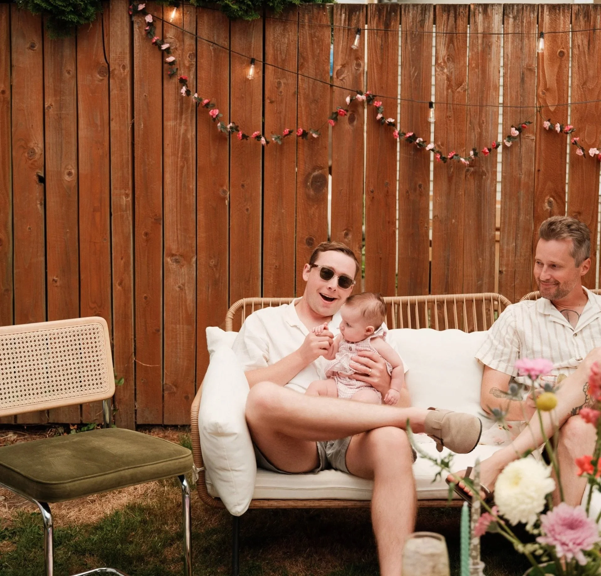 A group of people sitting outdoors on a patio behind a wooden fence, with some string lights hanging above. One woman is holding a baby, wearing sunglasses and laughing, while a man with a beard and short hair is sitting beside her, smiling. There ar