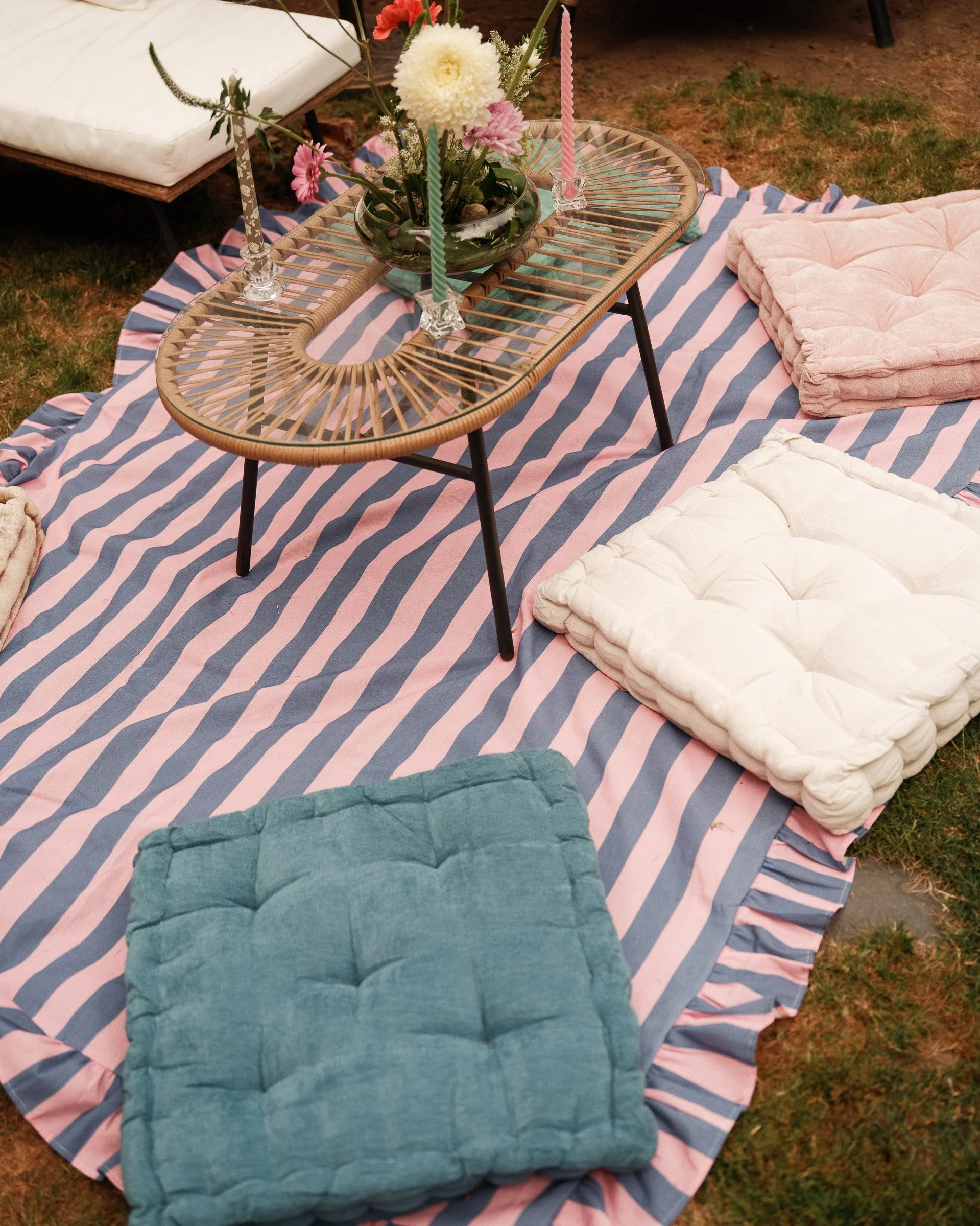 An outdoor picnic setup on the ground features a large striped pink and blue blanket. There are two stuffed cushions, one green and one cream, placed on the blanket. A wicker table with three colorful candles and a floral centerpiece with white and p