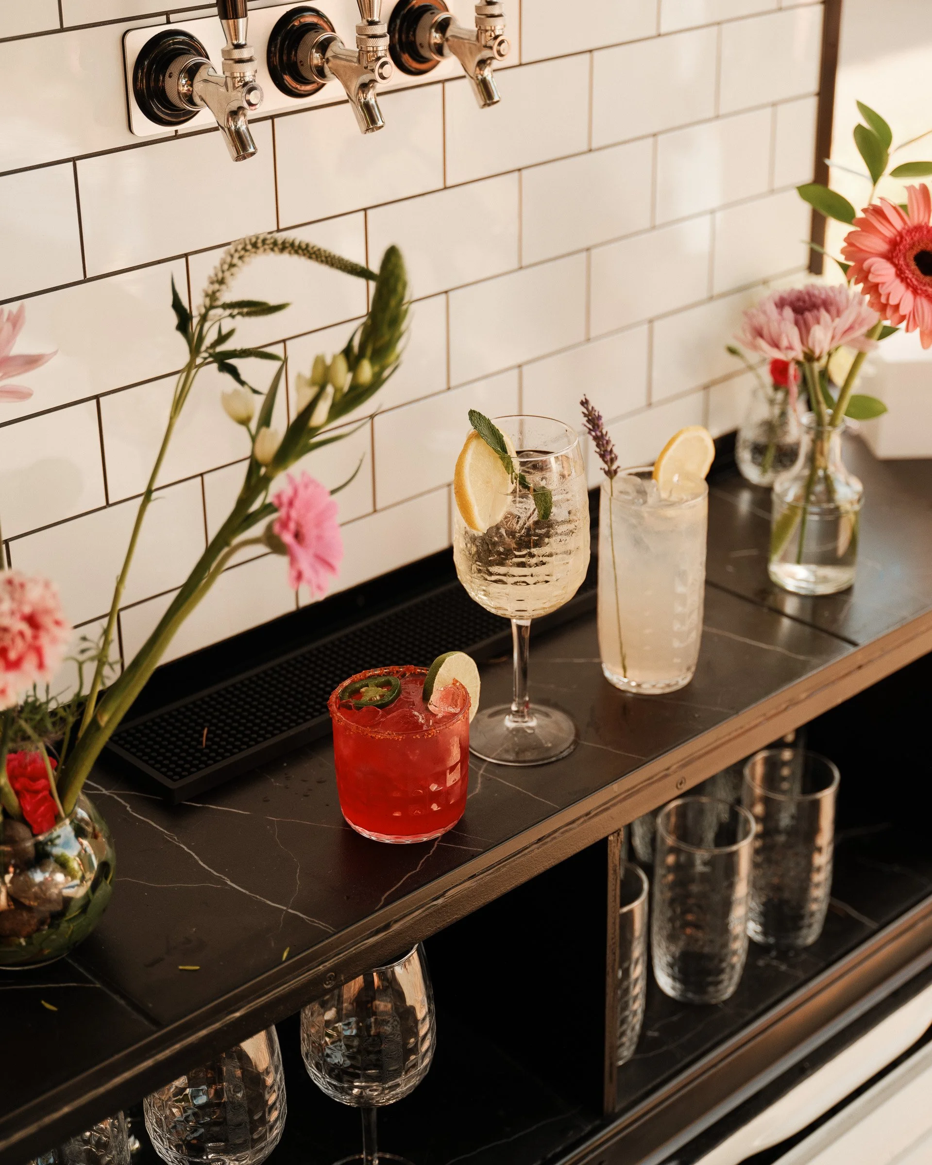 Close-up of a bar counter with floral arrangements and drinks, with a black tiled backsplash, brass beer taps, and empty glassware underneath.