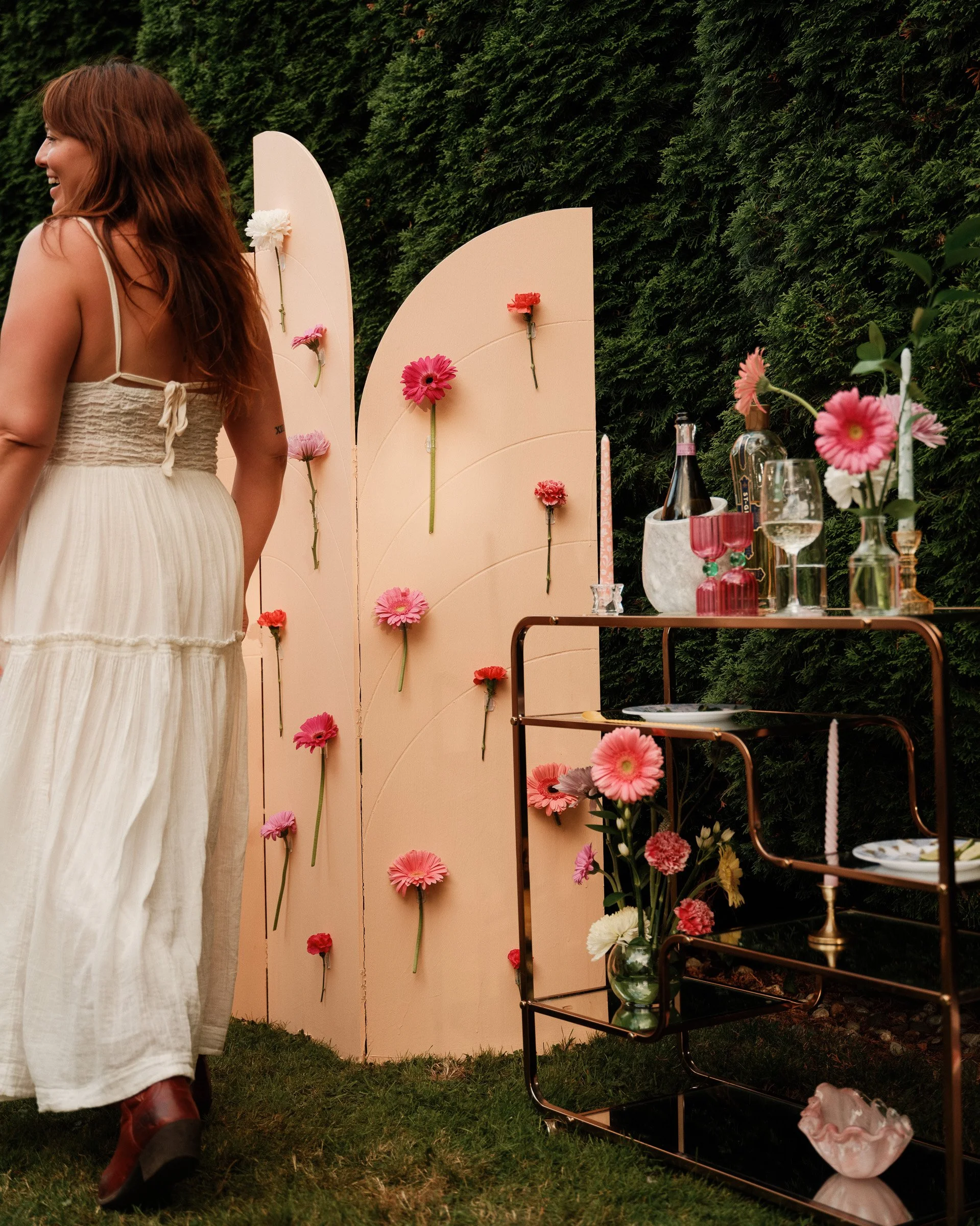 A woman with long red hair wearing a white dress and red boots standing on green grass at an outdoor event with floral decorations and a peach-colored folding screen adorned with colorful flowers, and a table with glassware, candles, and a bottle of 