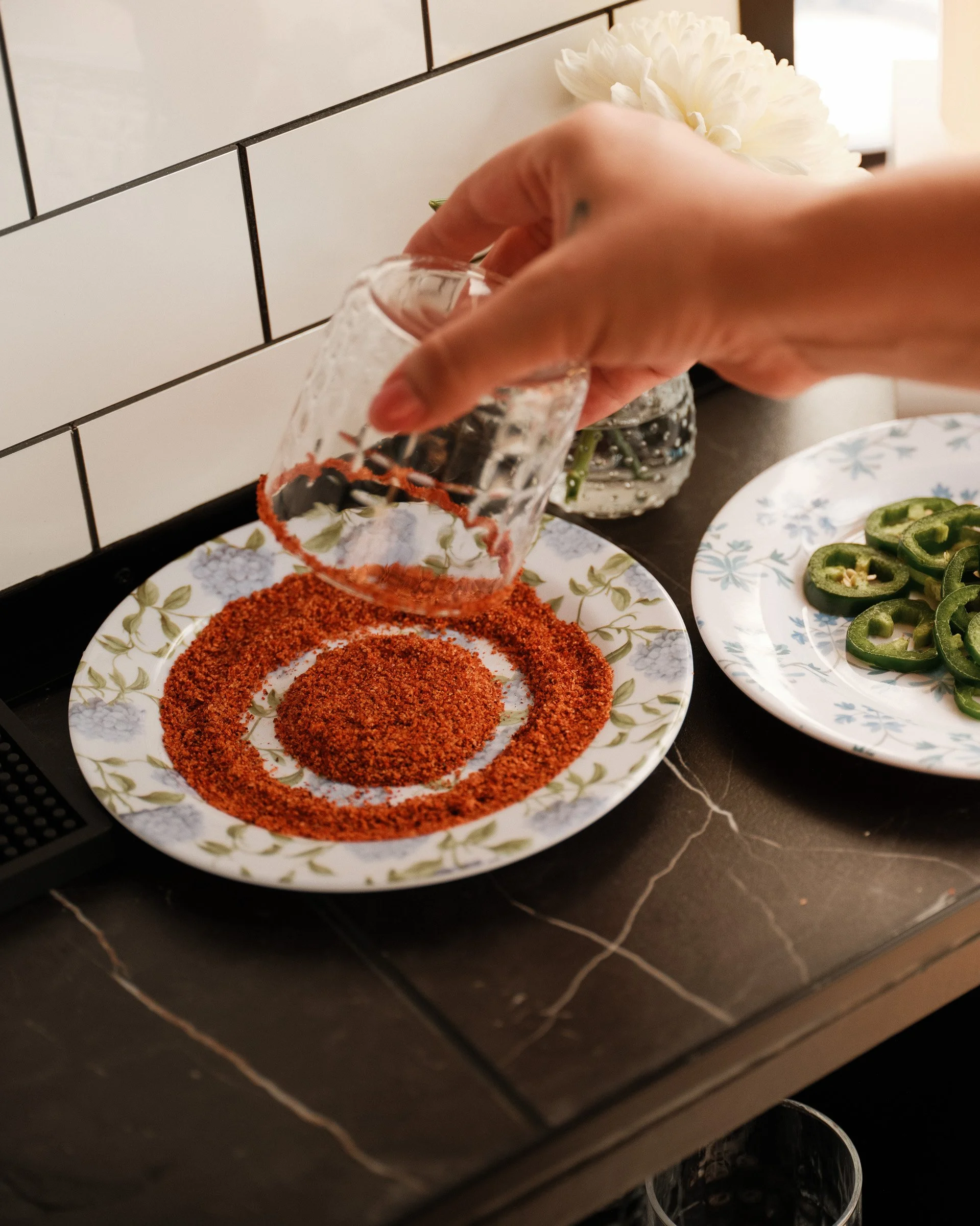 A person is pouring red chili powder onto a decorative plate with a floral pattern. There is a bowl of sliced jalapeño peppers and a white flower in a vase in the background, on a black kitchen counter with white-tiled backsplash.