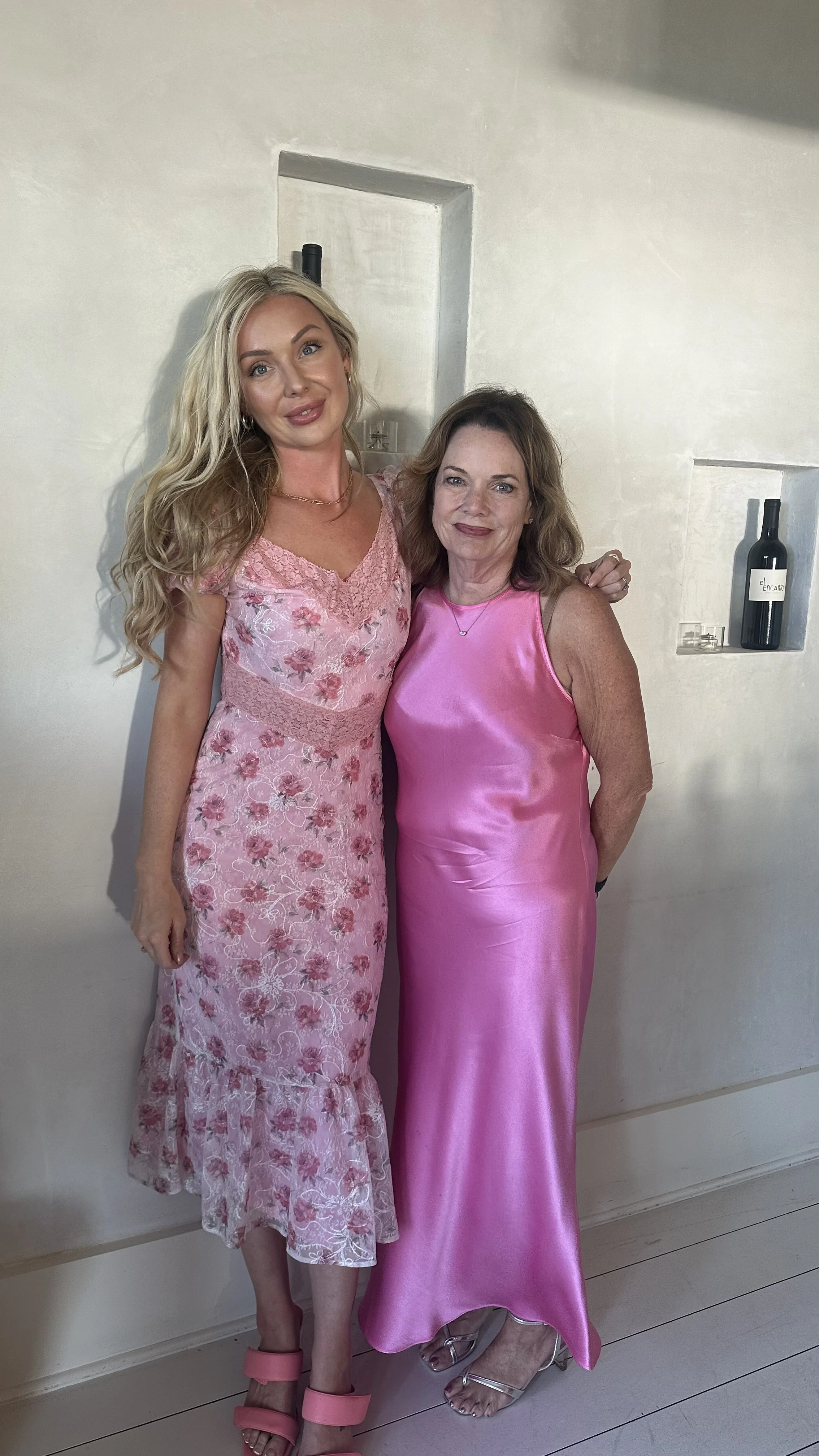 Two women standing close together, dressed in pink clothing, smiling in front of a white wall with built-in shelves.