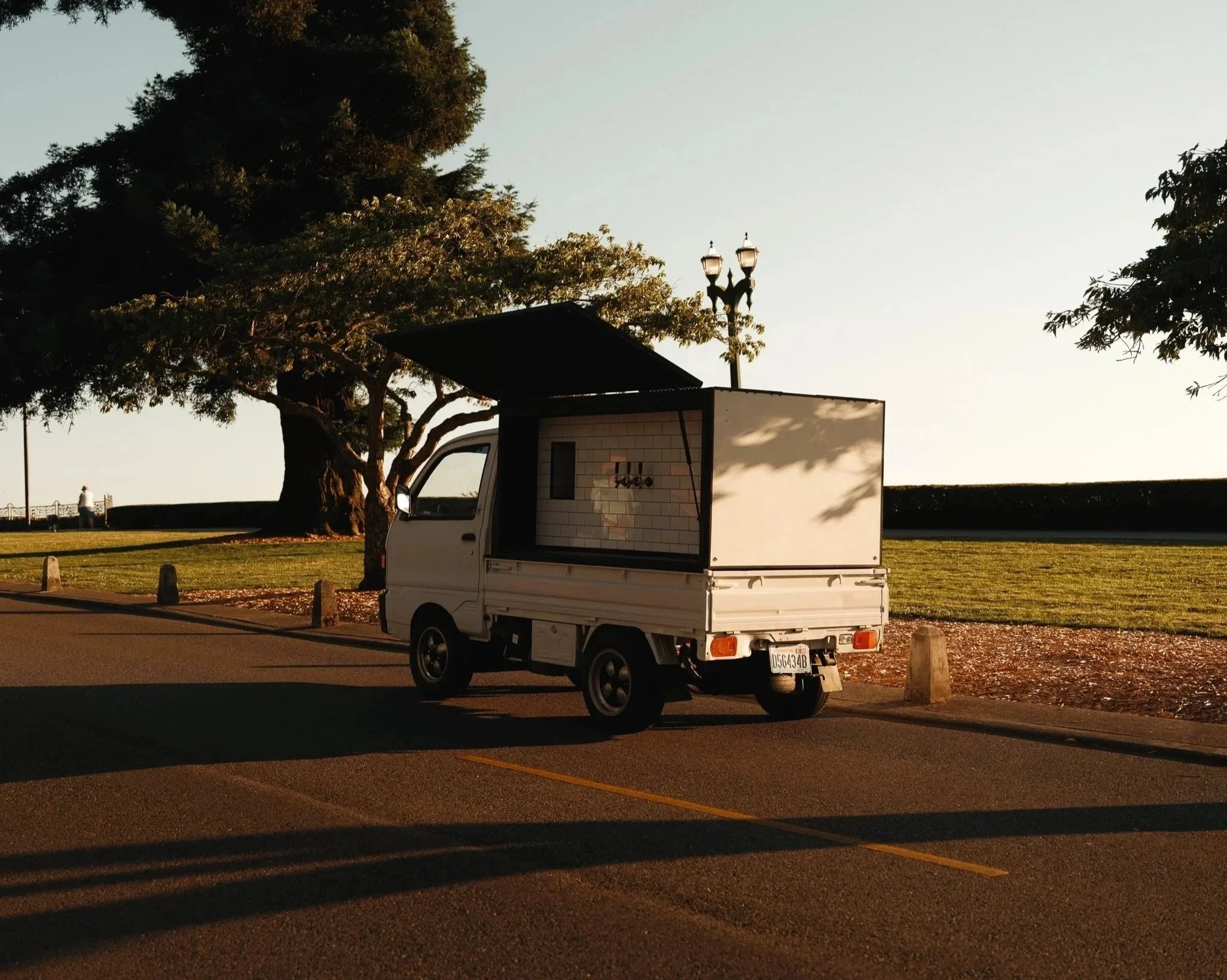 A small white delivery truck parked in an empty parking lot with a backdrop of trees. The truck's cargo area is open, revealing a tiled interior. In the background, there are a couple of people near a railing and a lamppost with multiple lights.