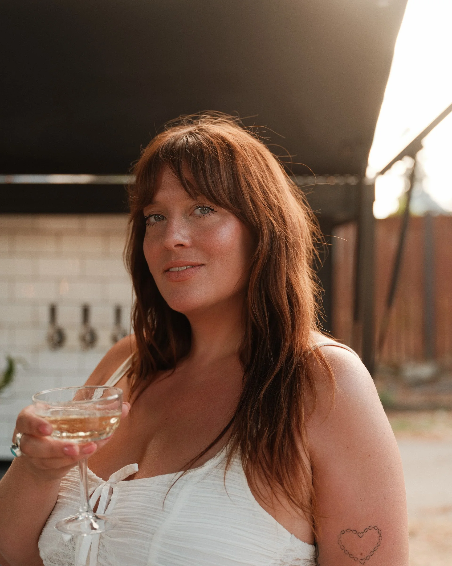 A woman with reddish-brown hair holding a glass of water, wearing a white top with lace, and a heart-shaped tattoo on her arm, standing outdoors during daylight.