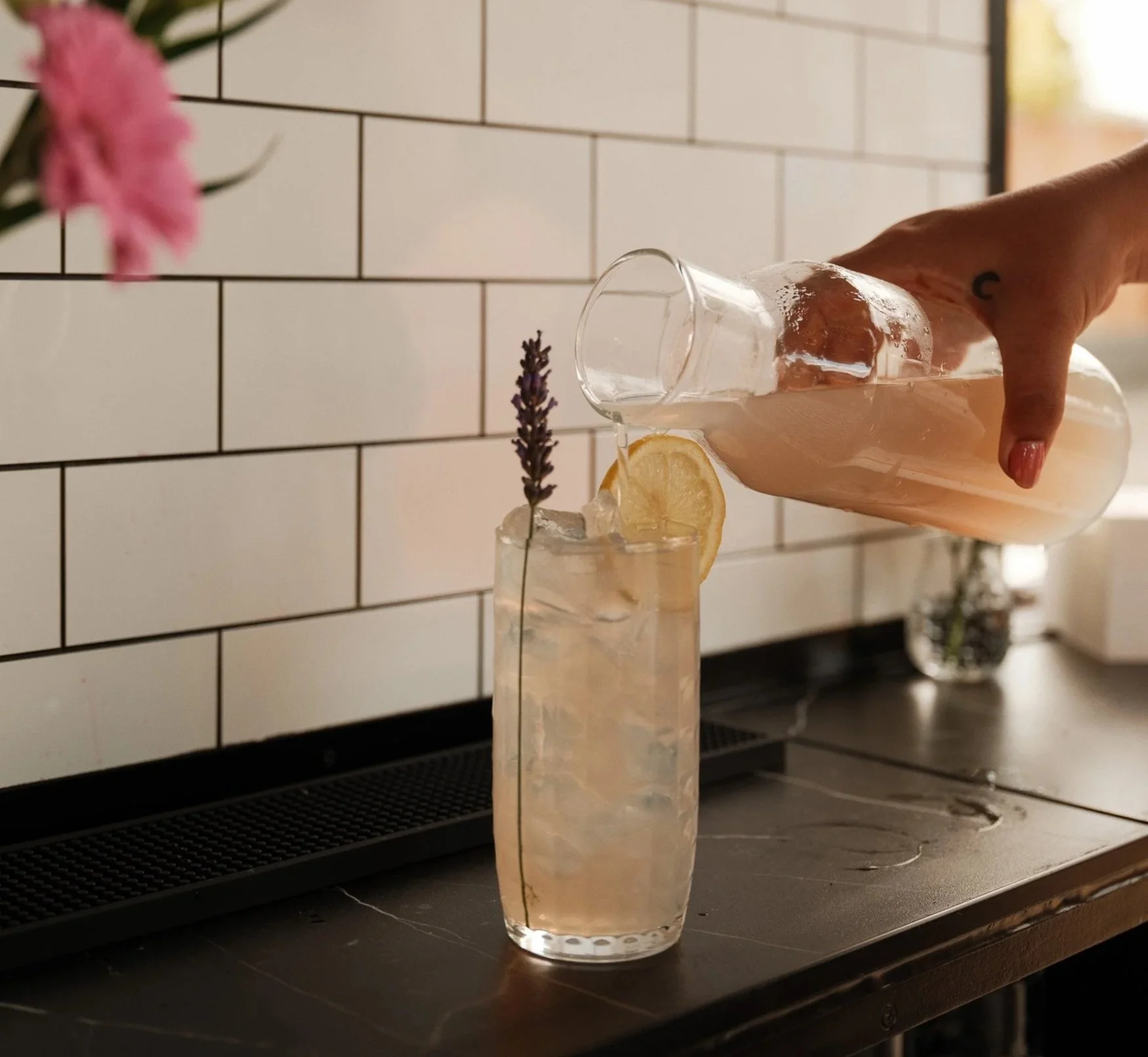 A person is pouring a pink cocktail from a glass bottle into a tall glass filled with ice, lemon slices, and a sprig of lavender, with a white tiled wall in the background.