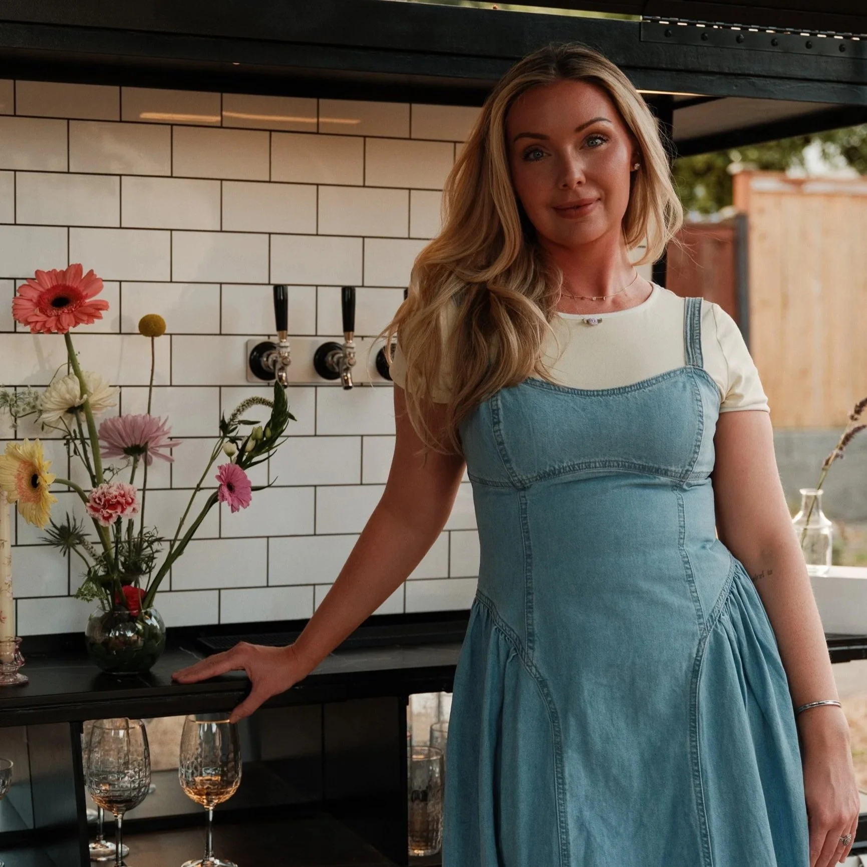 A woman with long blonde hair, wearing a light-colored shirt and denim dress, standing indoors by a black table with a vase of flowers and glasses of wine, with a white tiled wall and an outdoor setting in the background.