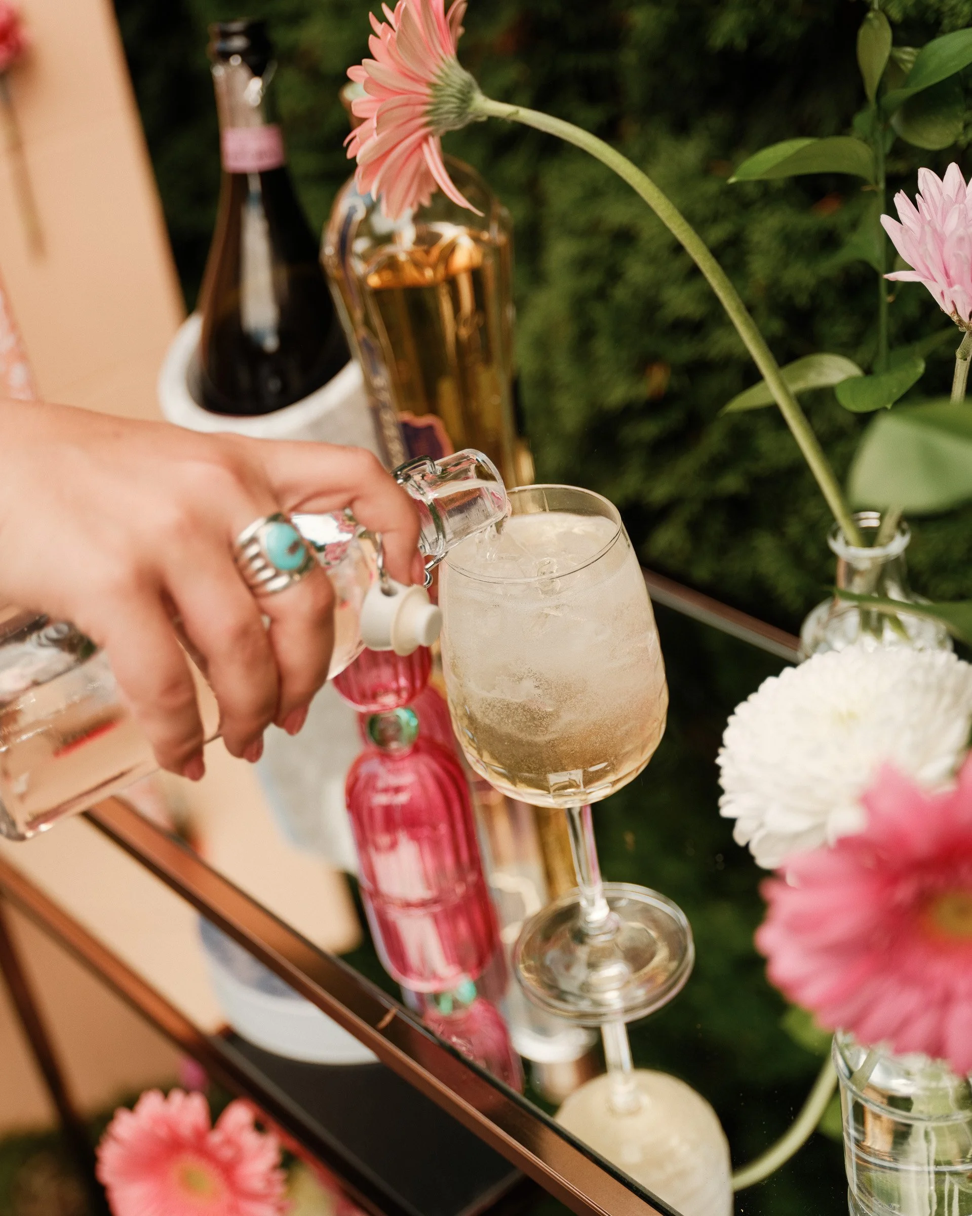 A person with rings on their fingers is pouring a drink into a glass with ice, near pink flowers and bottles, with a green leafy background.