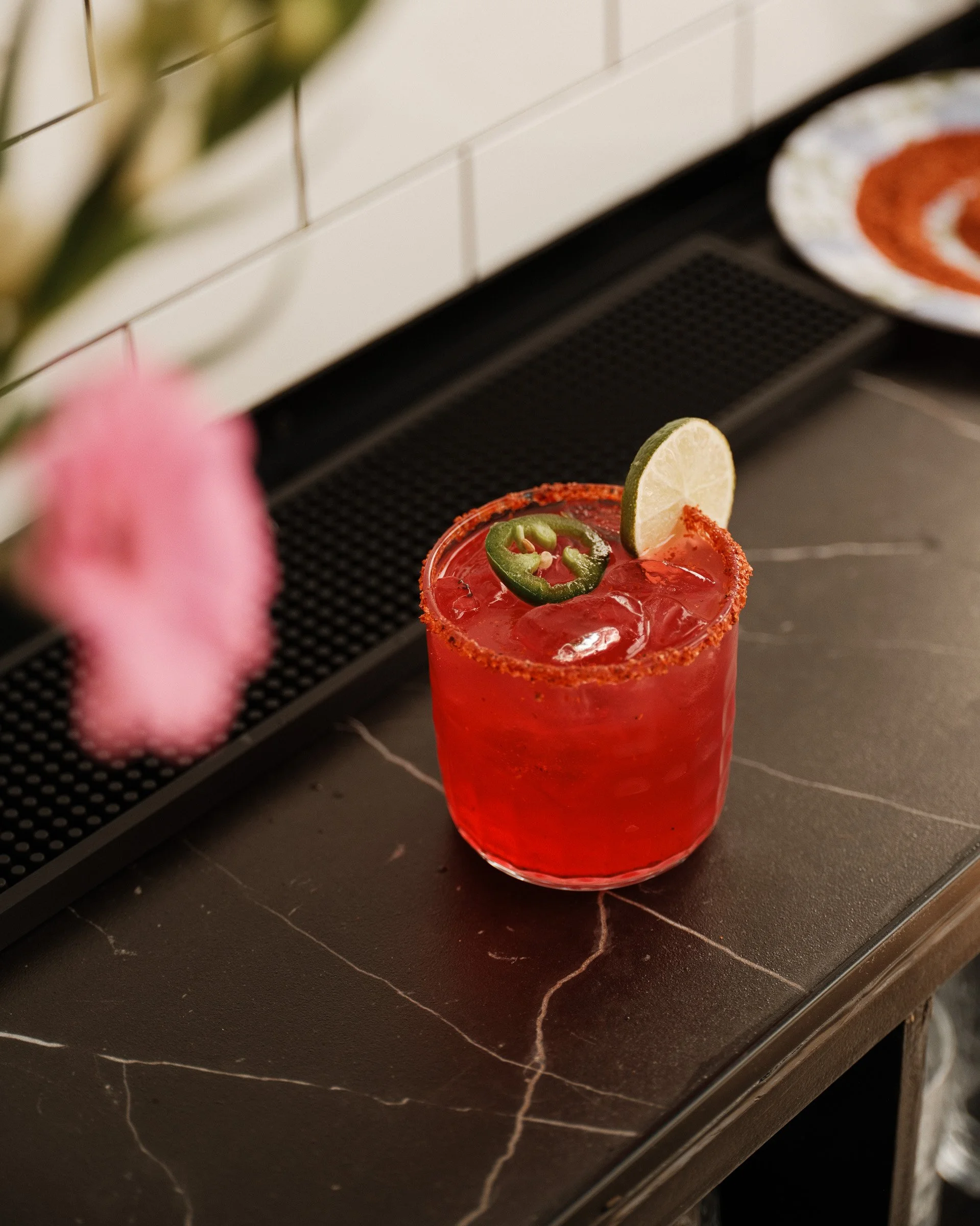 A close-up of a red cocktail with a salted rim, garnished with a lime wedge and sliced jalapeño pepper, placed on a black marble surface.