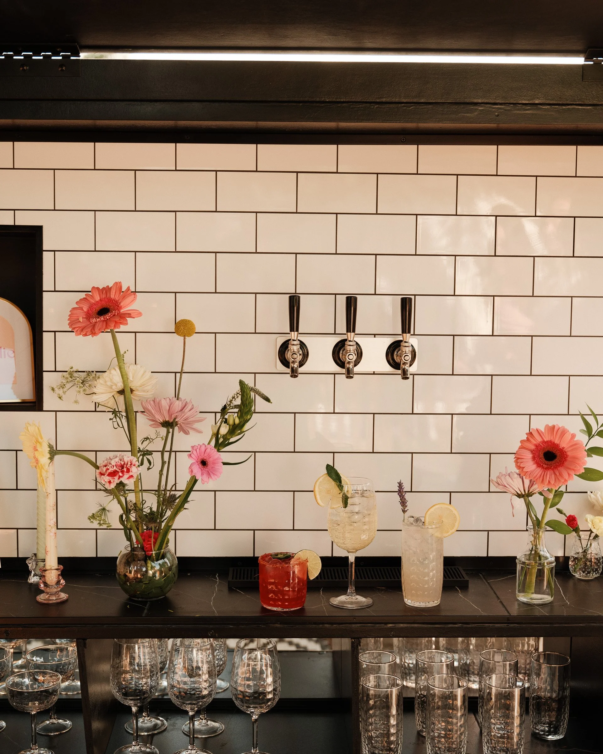 A bar display with various flowers in vases, cocktail glasses, and tumblers on a black countertop, set against a white subway-tile wall with alcohol taps.