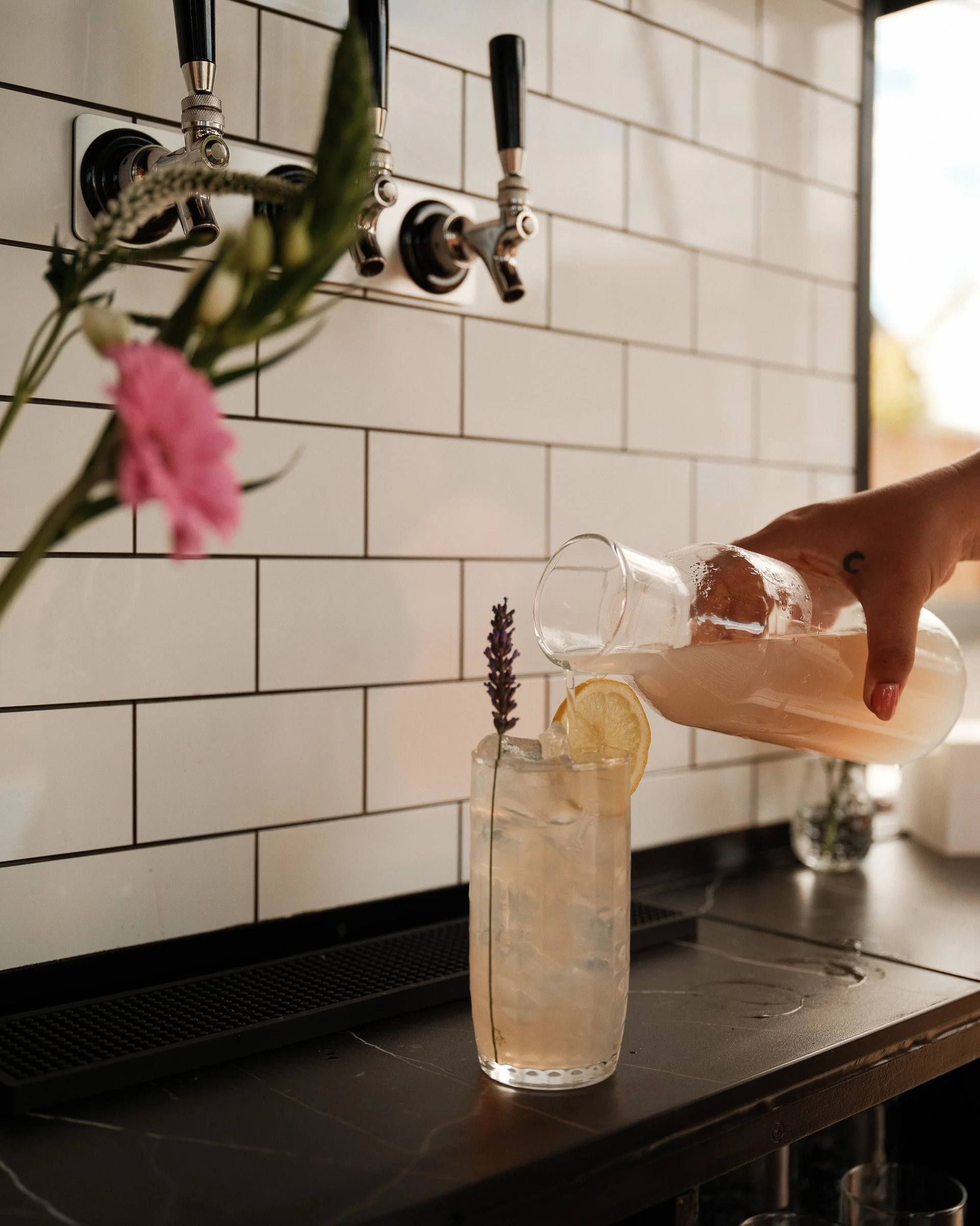 A tall glass of lemonade garnished with lemon slices and a sprig of lavender, placed on a black countertop with a white tiled wall background.