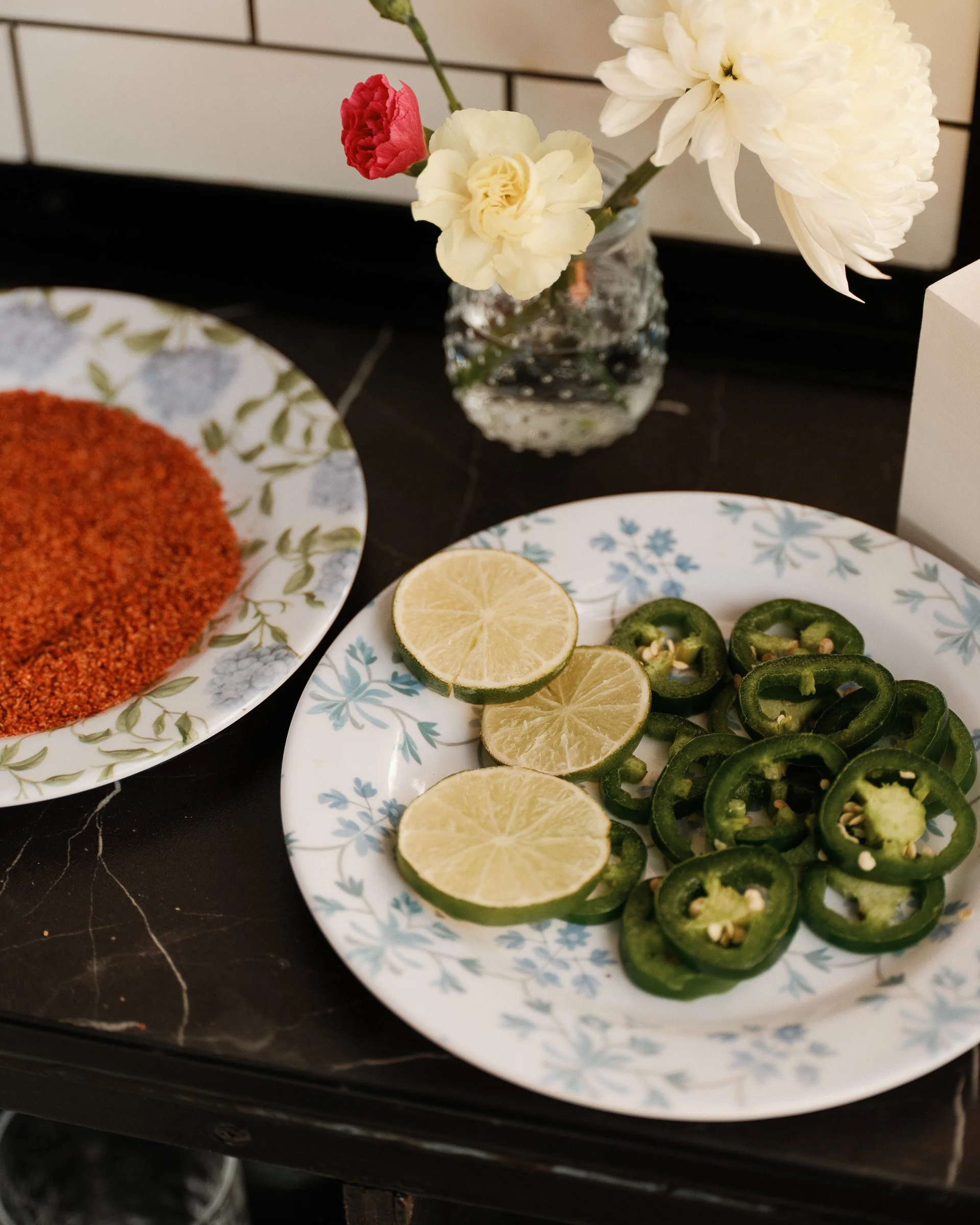 Close-up of sliced lemons and jalapeño peppers on a patterned plate, with a flower arrangement in the background.
