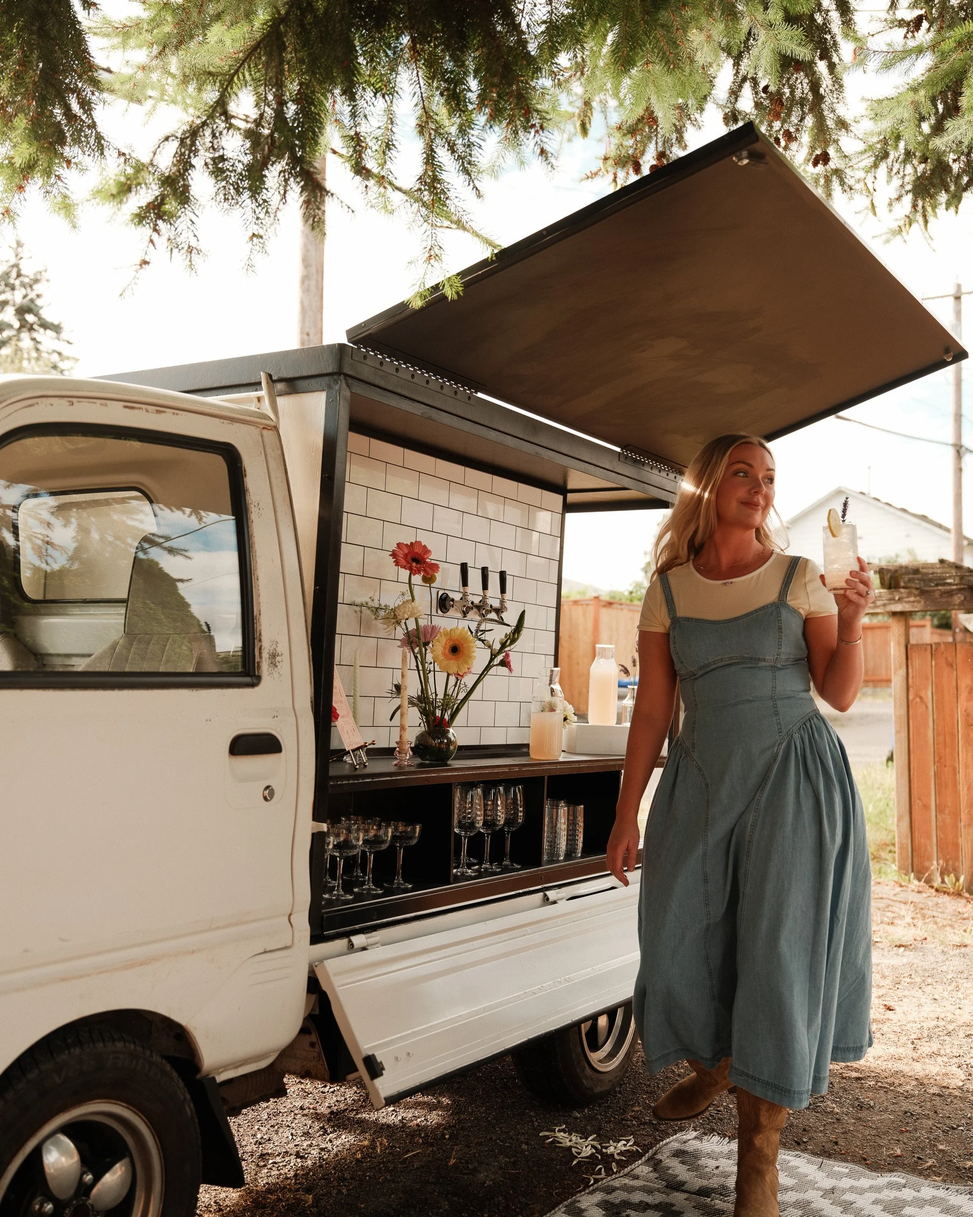 A woman with long blonde hair, wearing a denim jumper and boots, standing next to a mobile bar trailer with an open serving area. The trailer is white with a tiled front, decorated with flowers, and has glasses stored inside. The woman is holding a d