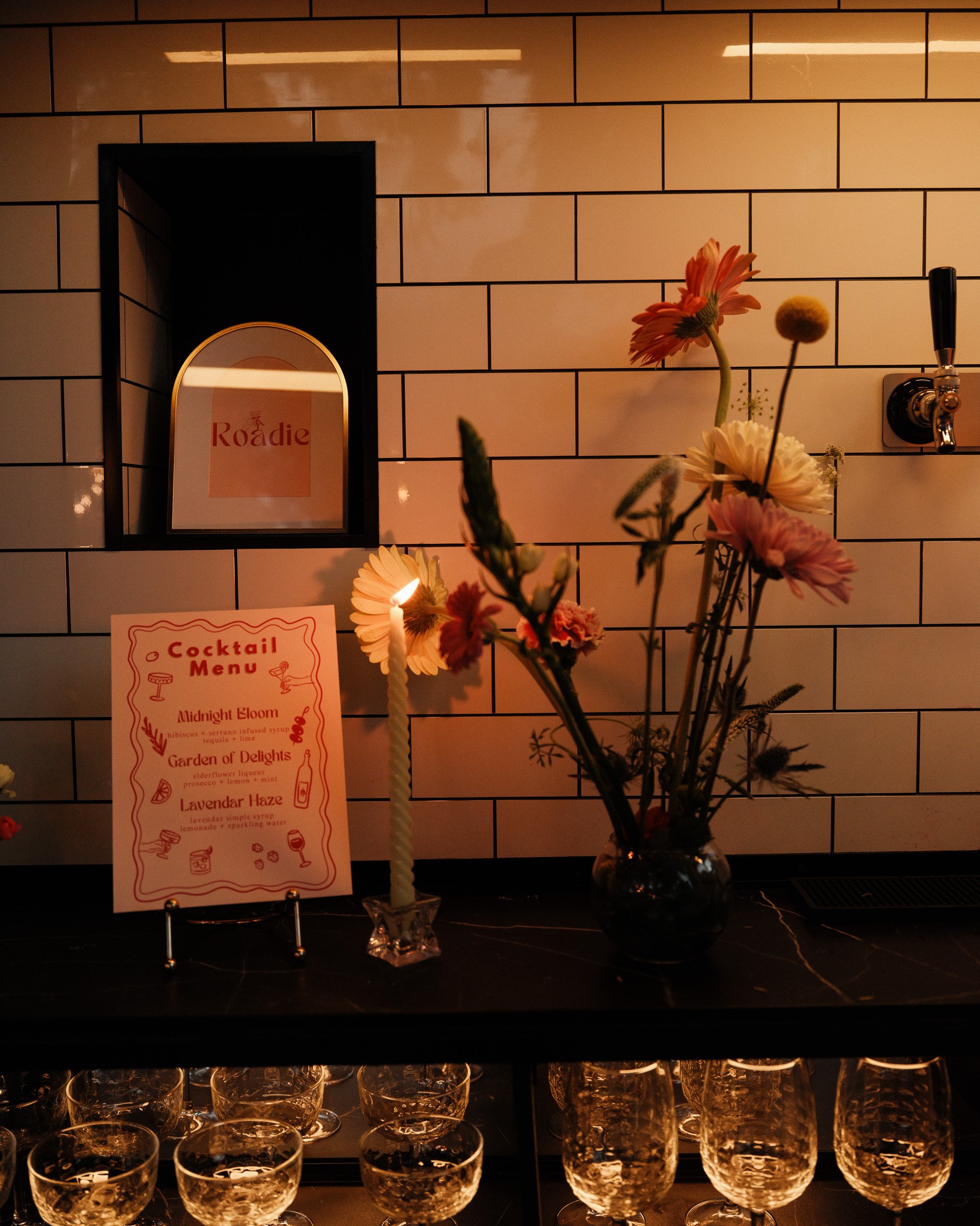 A bar setup with several empty glasses on a black counter, a glass vase with pink and white flowers, a sign for a cocktail menu, and a mirror reflecting a sign with the word "Roadie" on a bar tiled wall background.